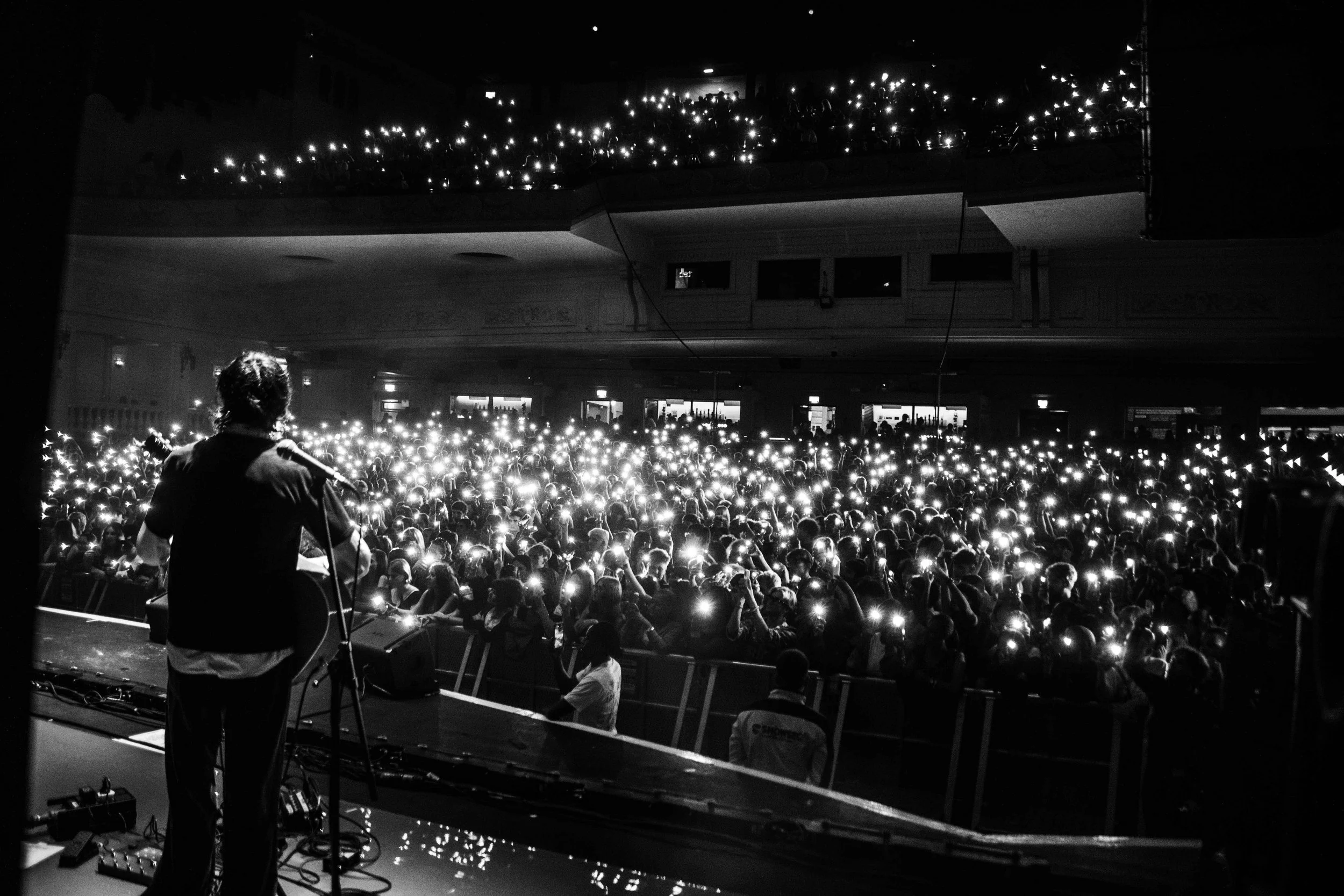 A performer with a guitar on stage facing a large audience in a concert hall, with the audience holding up their phones with lights on, creating a sparkling effect in the dark atmosphere.