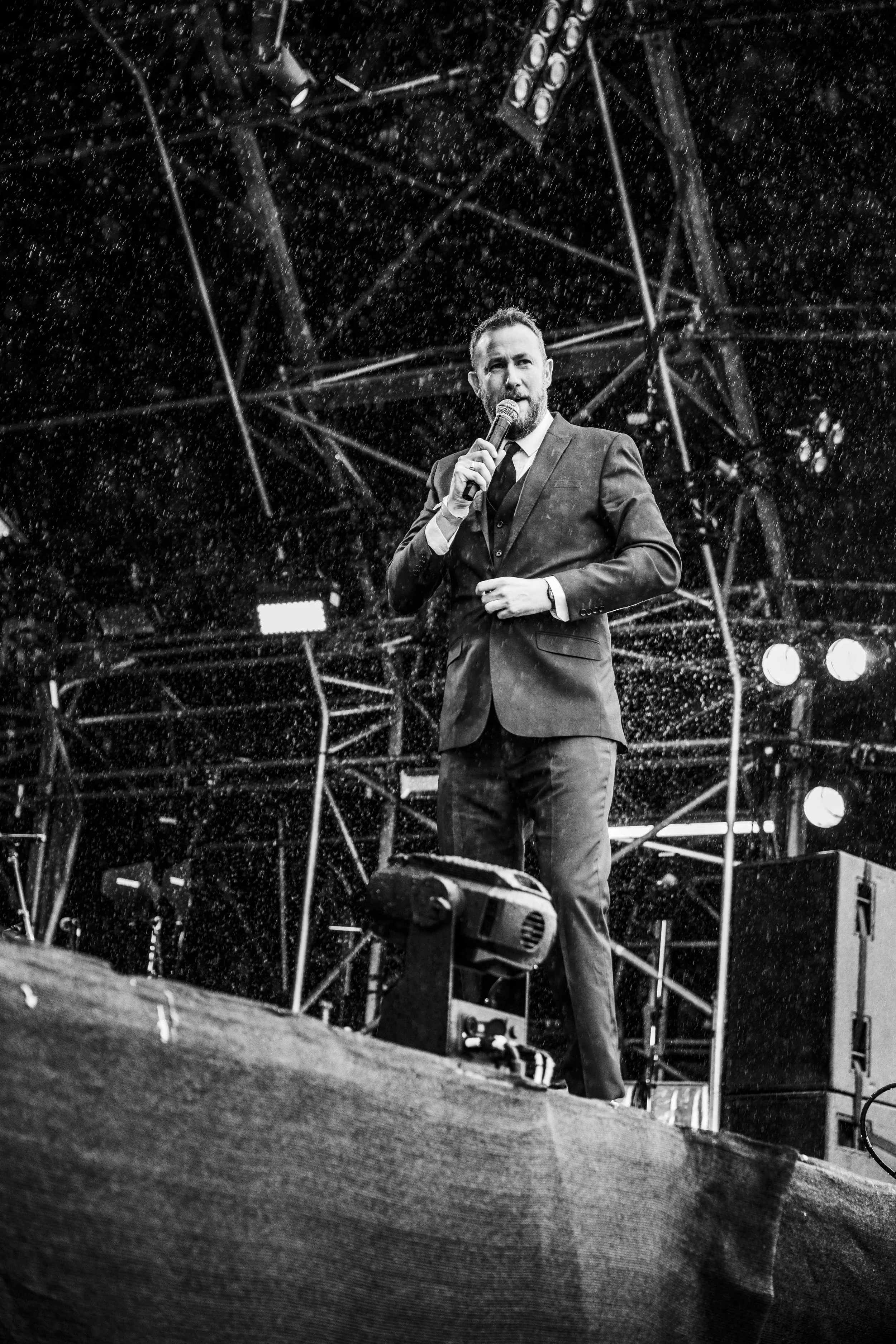 Man in suit speaking into microphone on stage in the rain, with stage equipment and scaffolding in background.