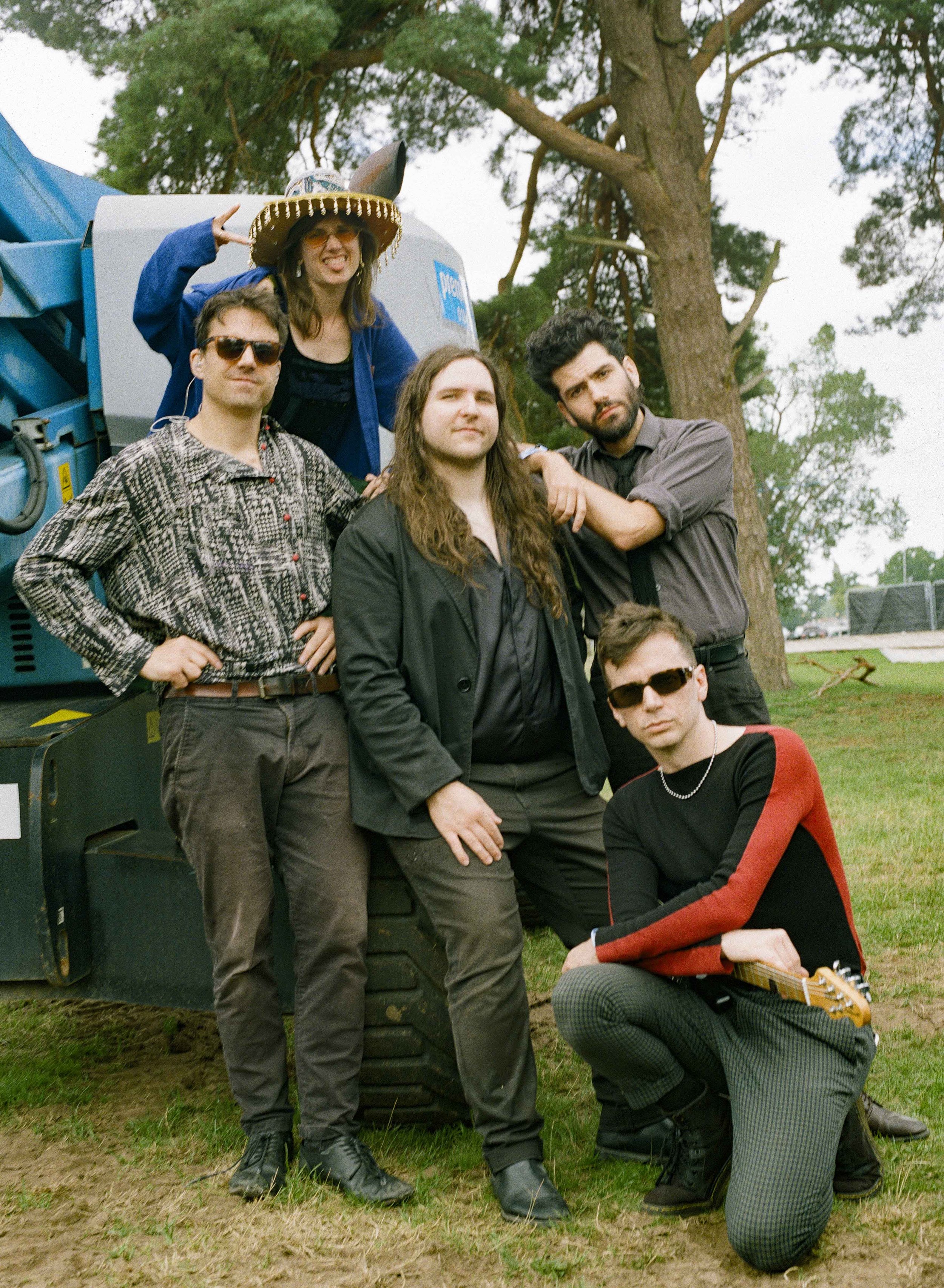 Group of five young adults outdoors in front of a tree and equipment, some wearing sunglasses, with one person wearing a sombrero hat and showing a peace sign.
