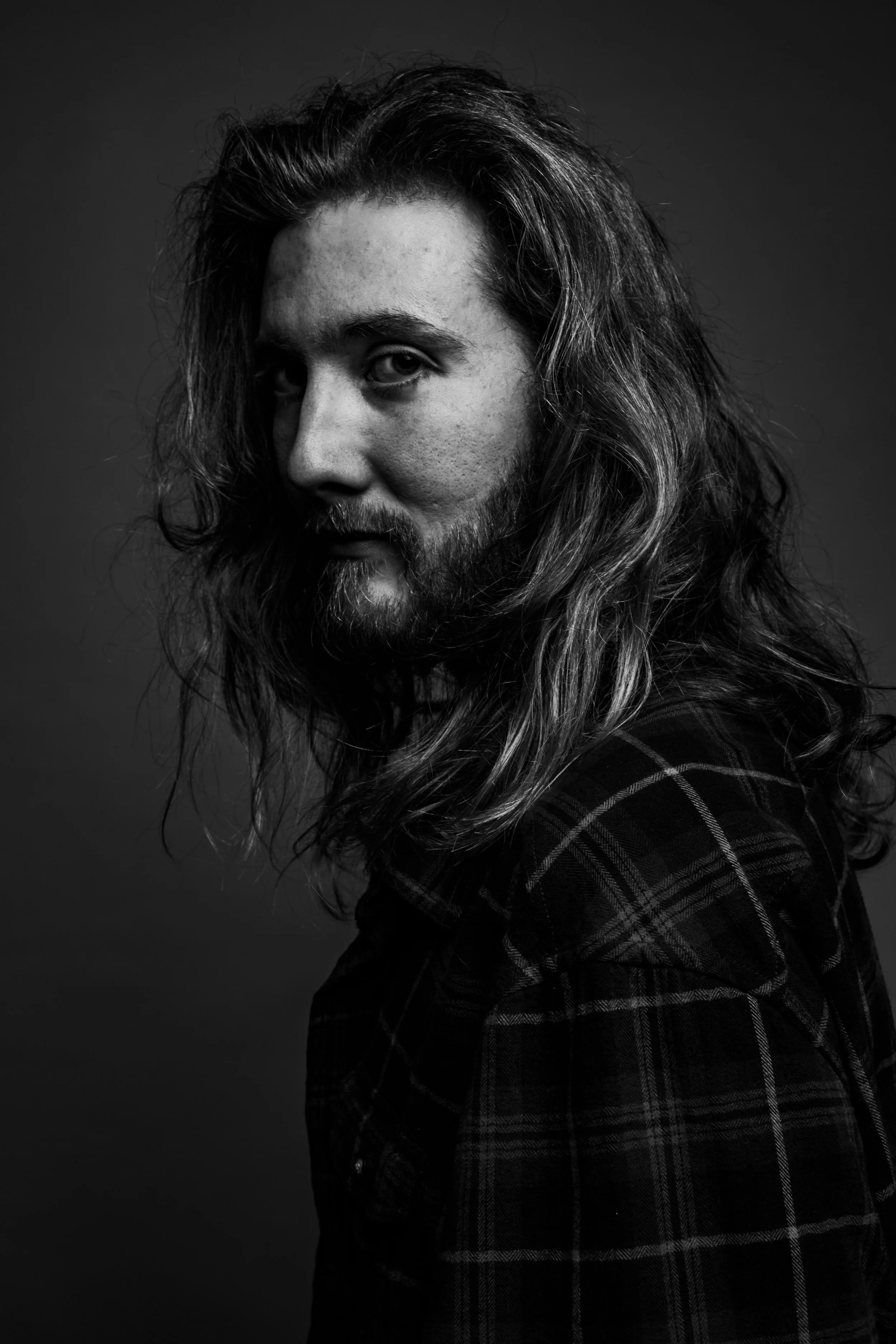 Black and white portrait of a man with long, wavy hair and a beard, wearing a plaid shirt.