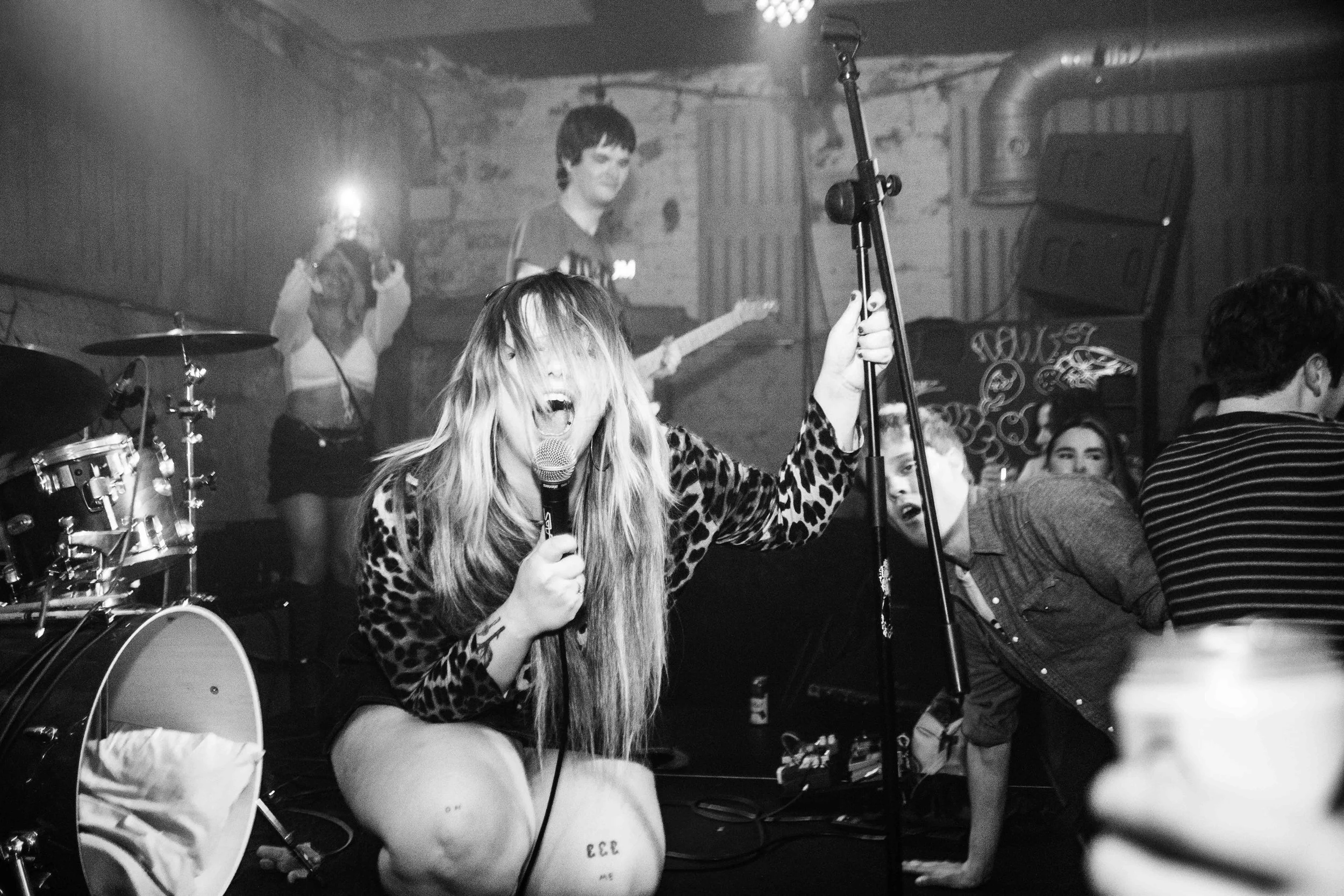 A woman singing into a microphone with long hair covering her face, holding a stand with her left hand, in a black-and-white photo at a live music venue. Band members are playing instruments in the background, and people are watching the performance.