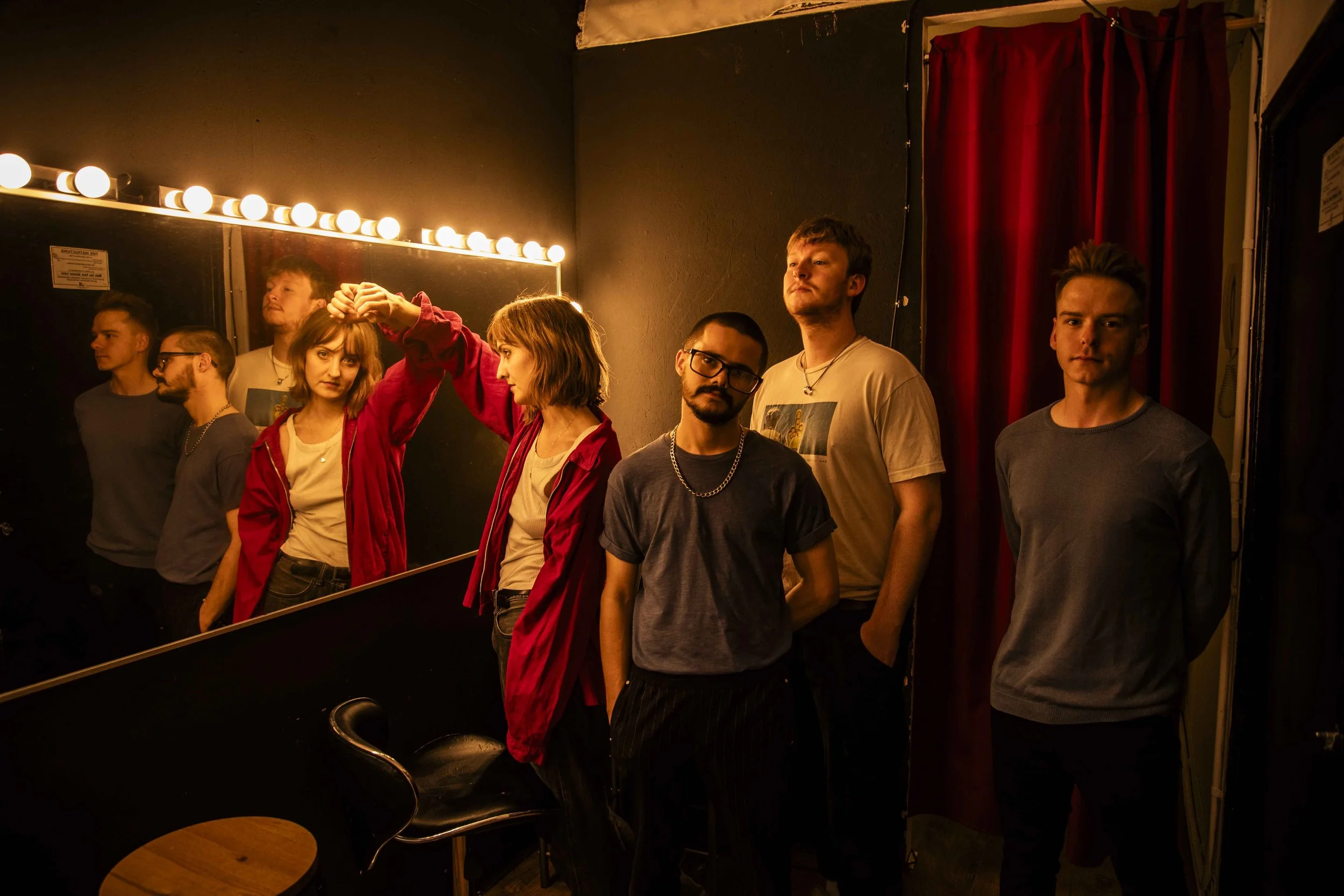 A group of seven young adults standing in a room with a black wall and red curtains, with some looking into a mirror and others looking at the camera. One person is touching another's head, and a black chair and a round table are visible in the foreg