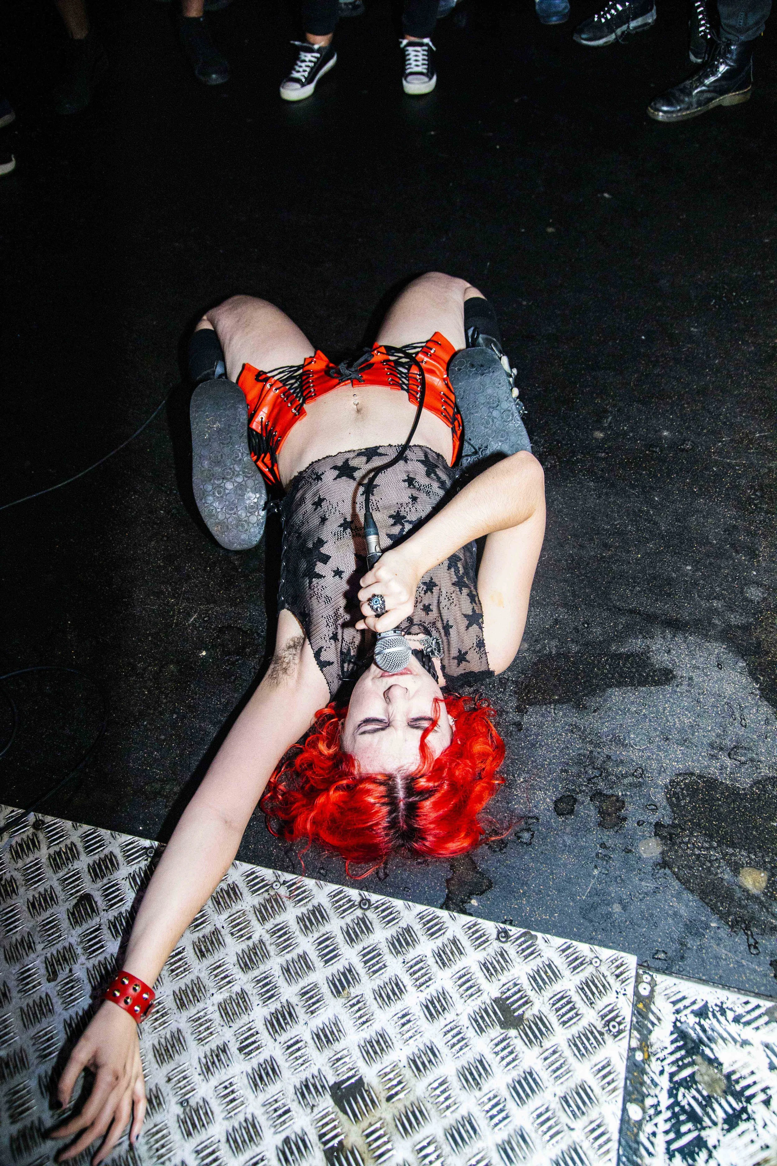 Performer with bright red curly hair lying on stage floor, holding microphone, eyes closed, wearing black lace top, red shorts with black laces, black boots, and a red bracelet, surrounded by standing audience.