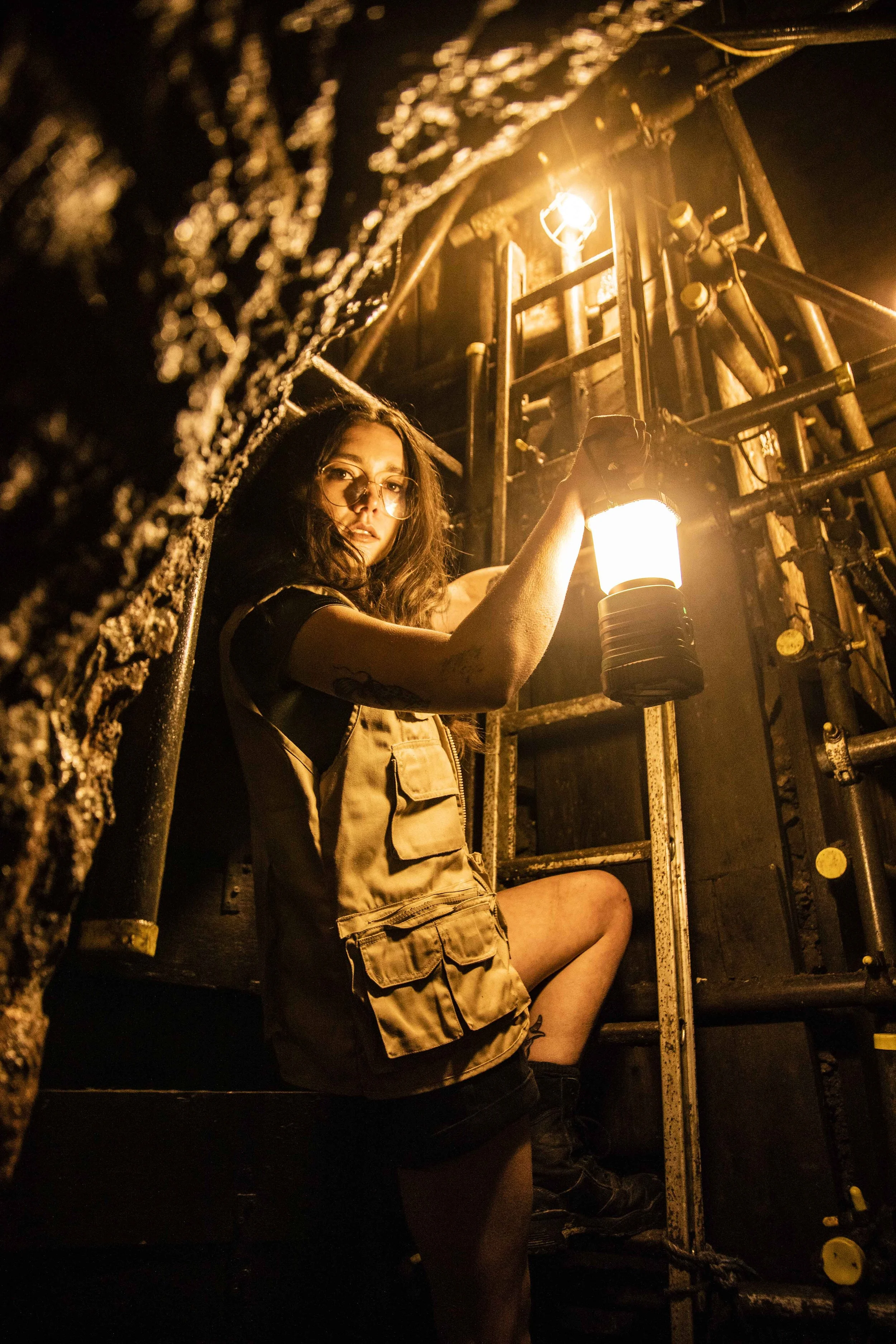 A young woman with glasses and tattoos holding a lantern while hanging from a ladder in a tunnel, illuminated by warm light from the lantern and nearby mining style light.