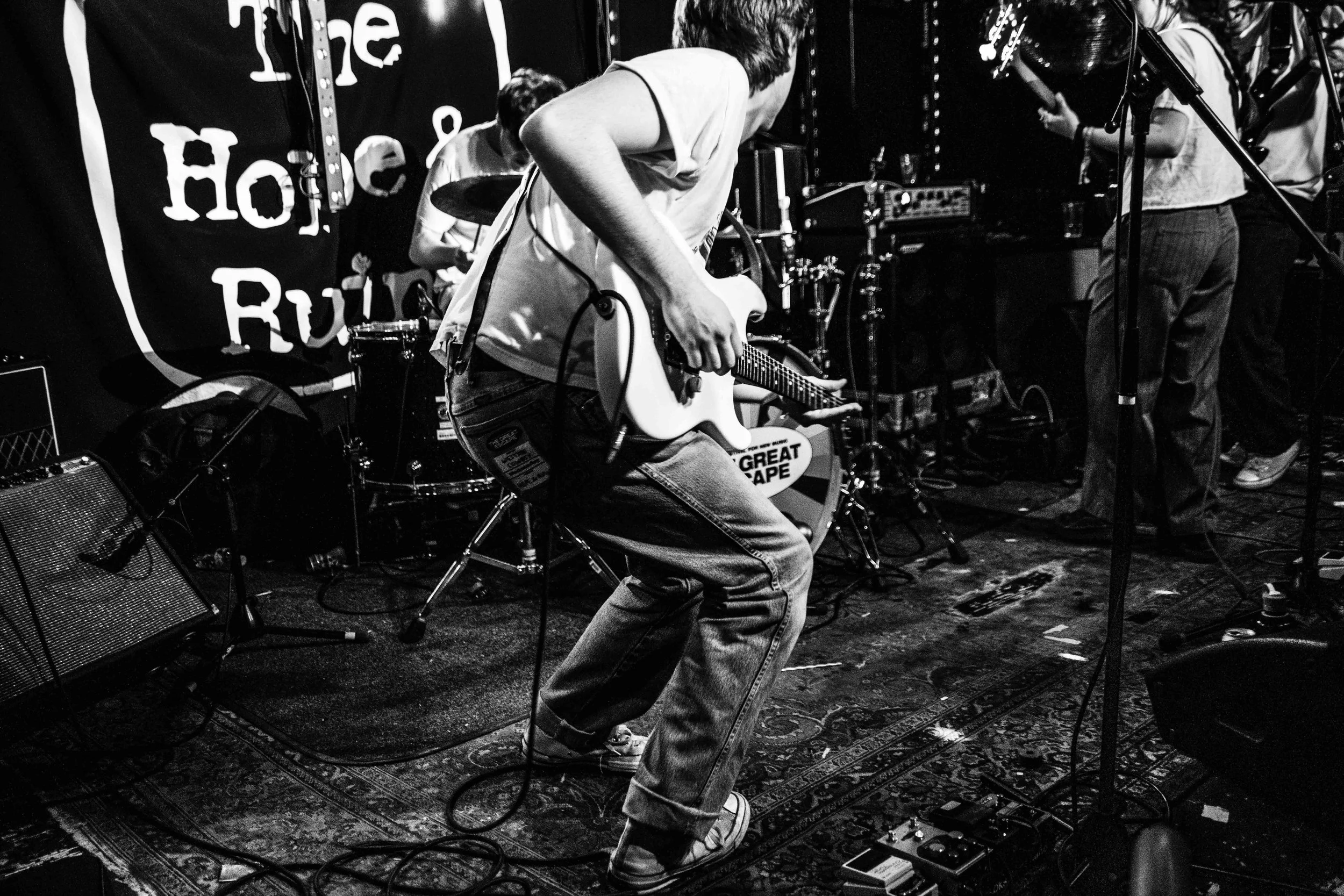 A black and white photo of a band performing on stage; the focus is on a person playing an electric guitar, bending forward, wearing a t-shirt and jeans. Other band members and stage equipment are visible in the background.