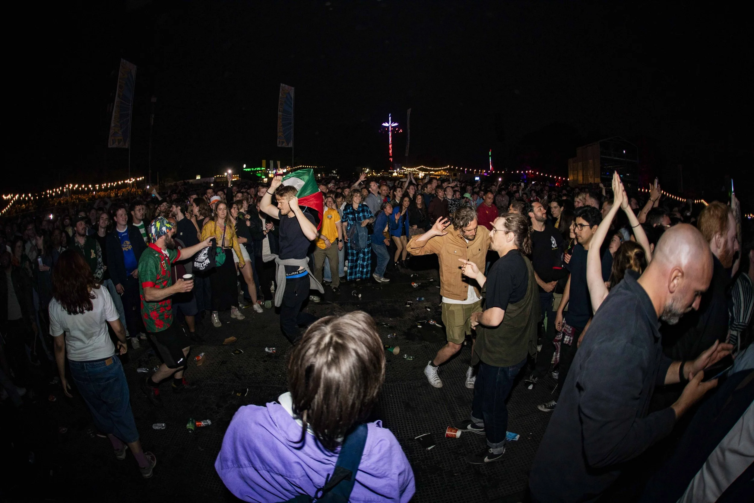 Crowd of people dancing and enjoying a nighttime outdoor event with colorful lights and fireworks in the background.
