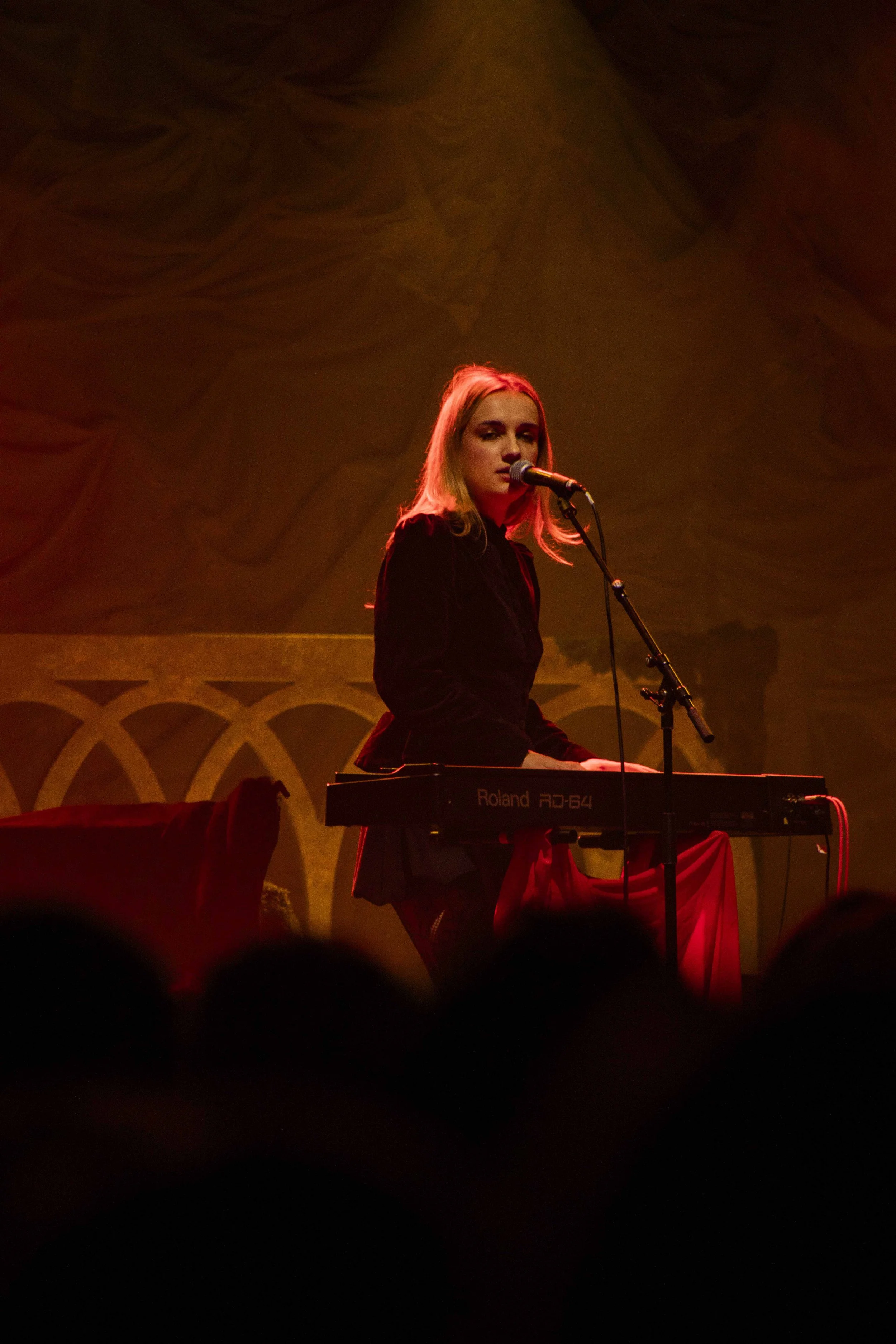 A woman with blonde hair wearing a black outfit, performing on stage playing a Roland RD-64 keyboard and singing into a microphone, with warm stage lighting and patterned backdrop.