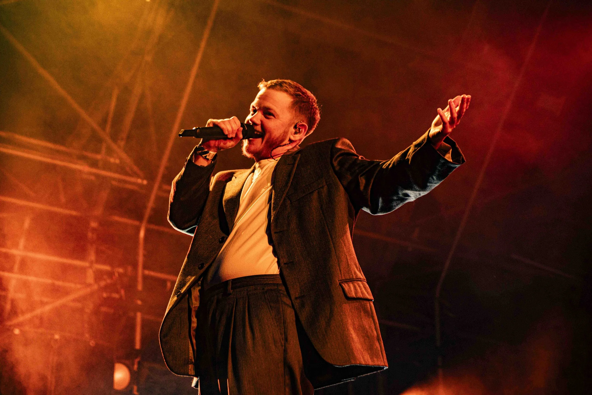 A masc appearing non-binary rapper performing on stage with a microphone, wearing a suit, bathed in warm stage lighting.