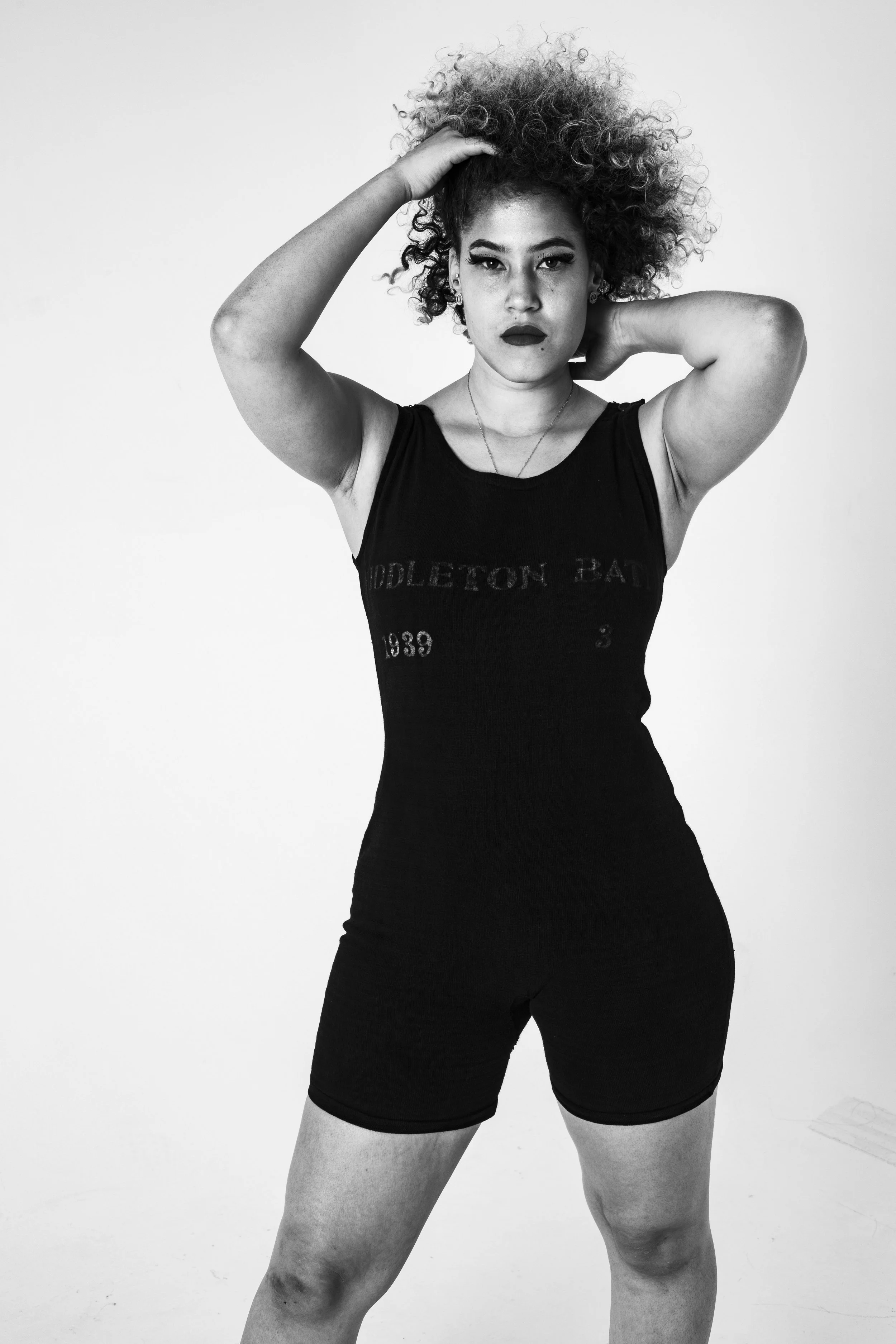 Black and white photo of a woman with curly hair, wearing a black sleeveless swimming costume from 1935, posing with one hand on her head and the other behind her neck.