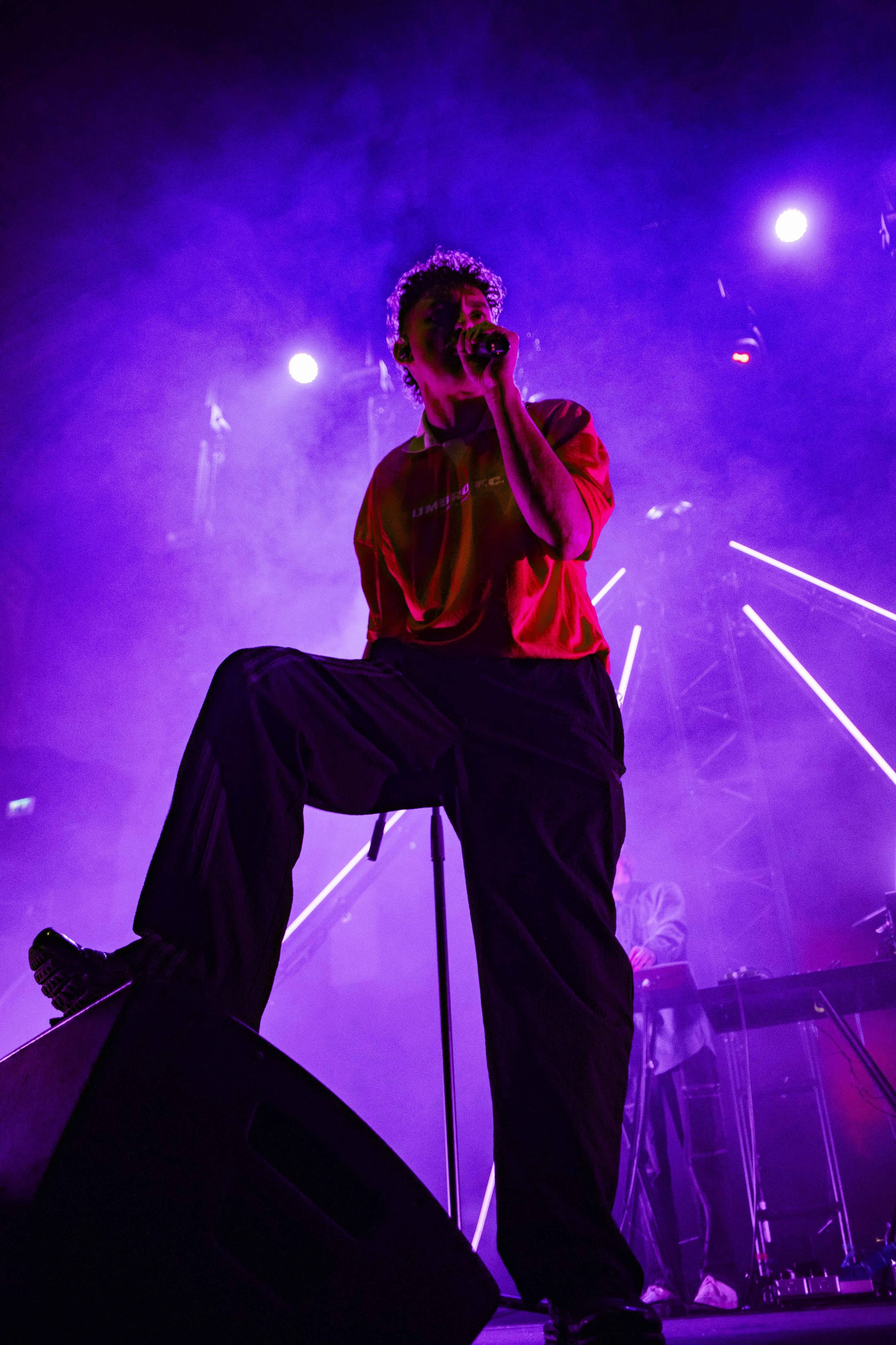 A male performer singing on stage with purple lighting and smoke effects, wearing a red shirt and black pants, while holding a microphone.