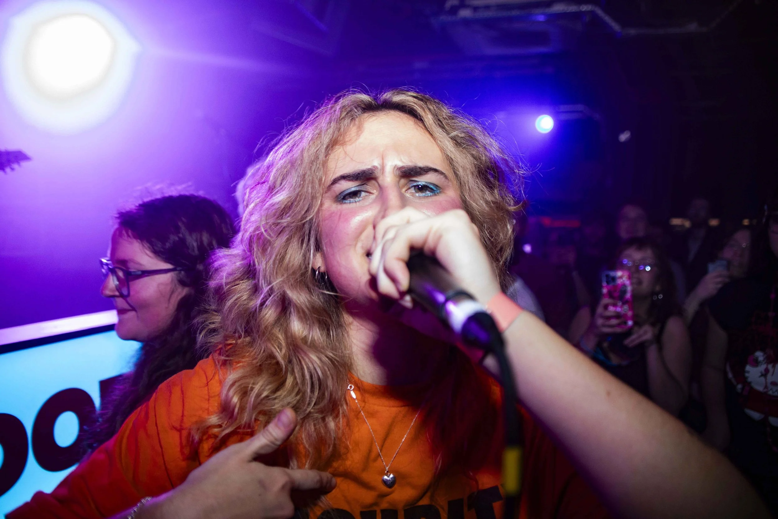 A woman with curly blonde hair singing into a microphone at a concert, with people in the background taking photos or watching.