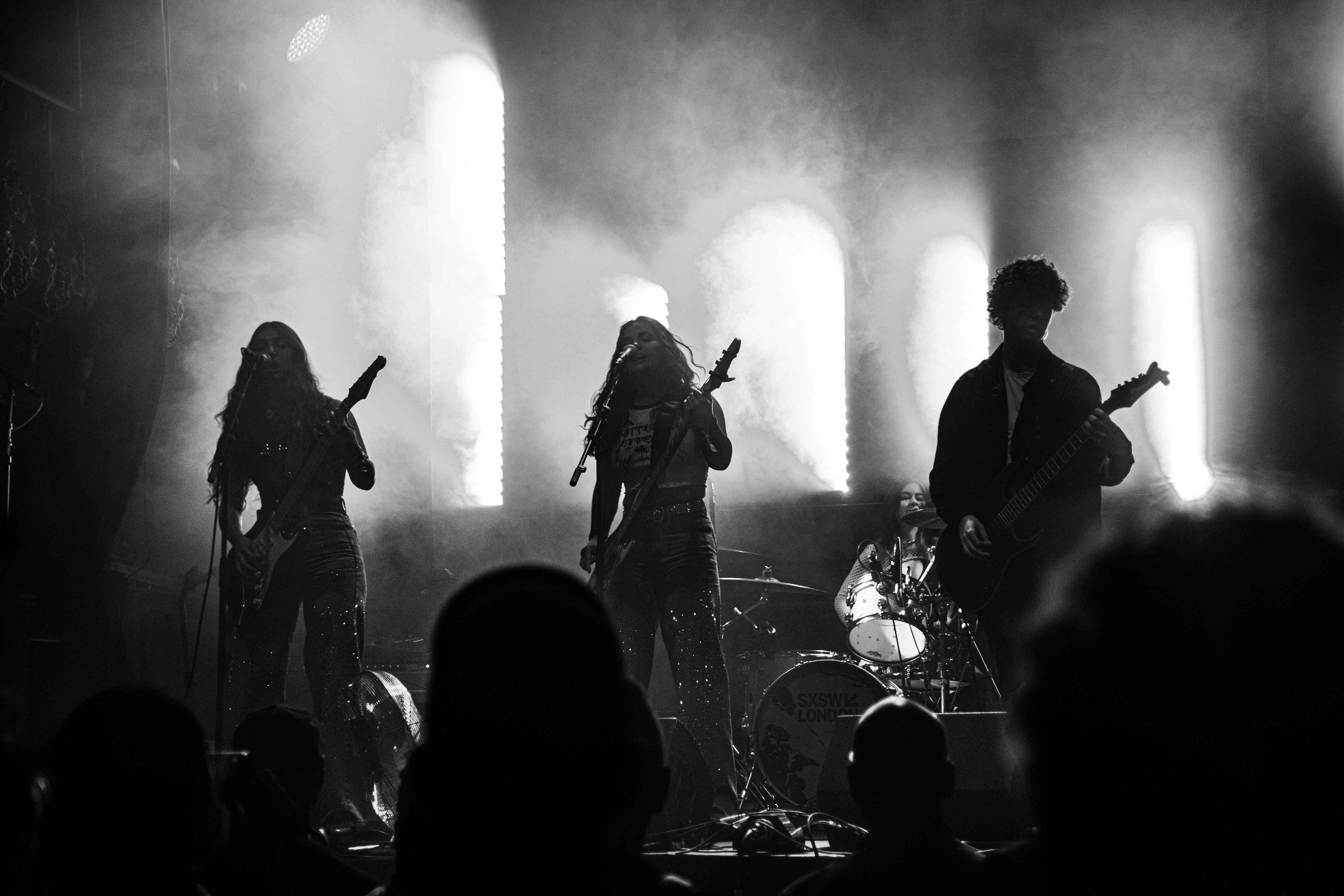 A black-and-white photo of a band performing live on stage, consisting of two female guitarists, a male guitarist, and a female drummer, with bright lights and smoke creating a dramatic atmosphere.