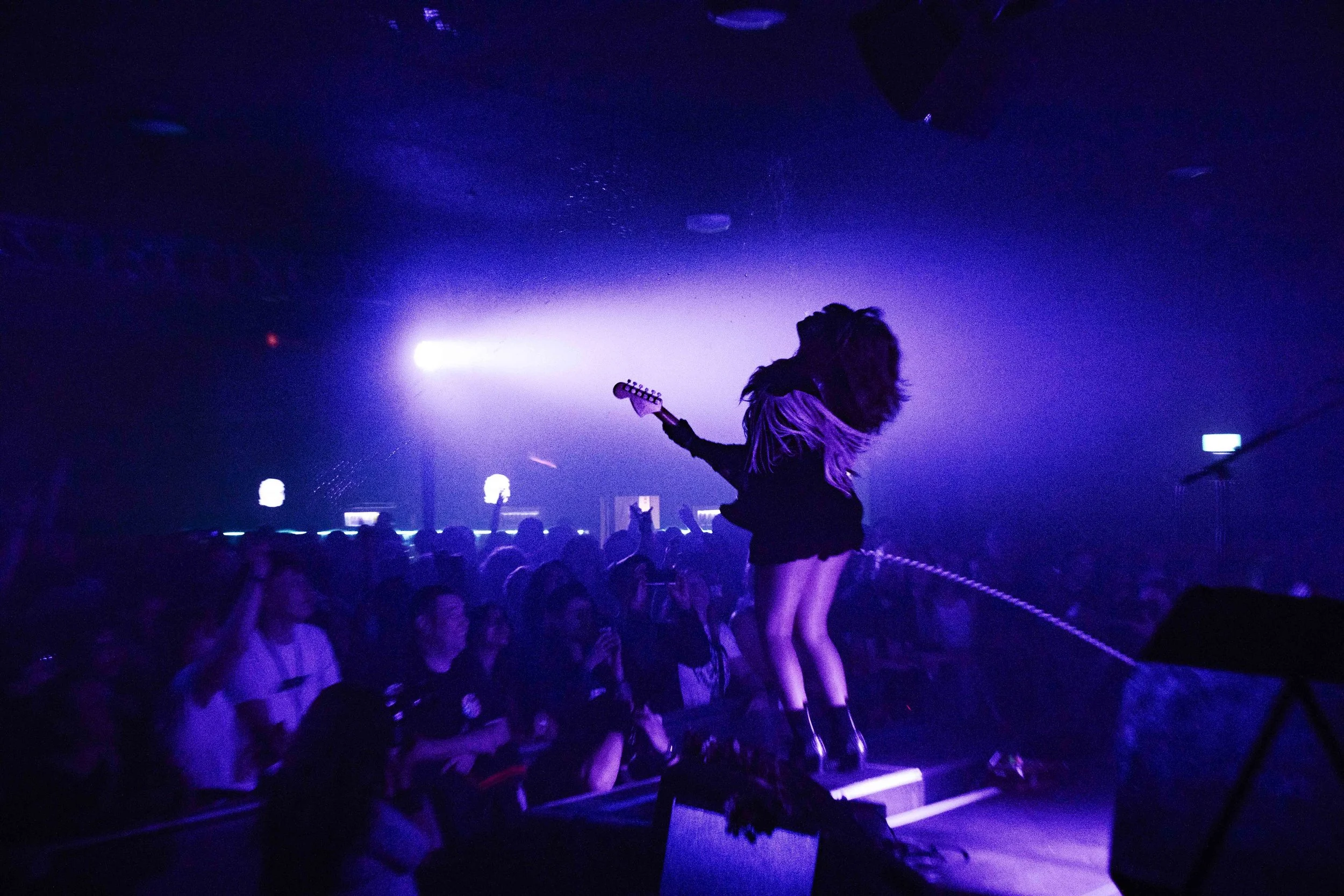 A female performer with long, wavy hair, dressed in black, stands on stage with a guitar, leaning back as she interacts with the audience in a dark, purple-lit concert venue filled with people.