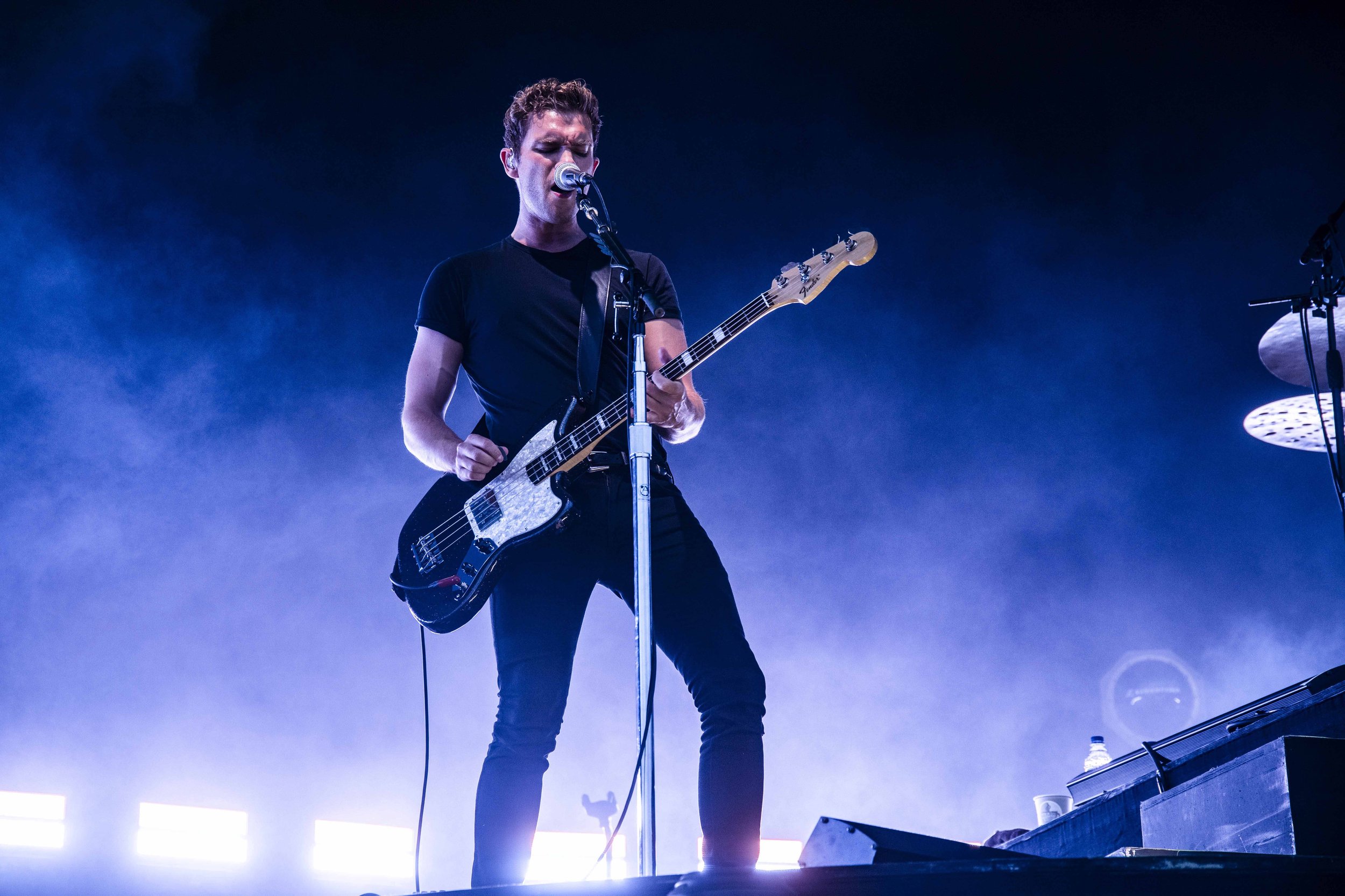 A musician performing on stage with a bass guitar, singing into a microphone, with blue stage lighting and fog effects.