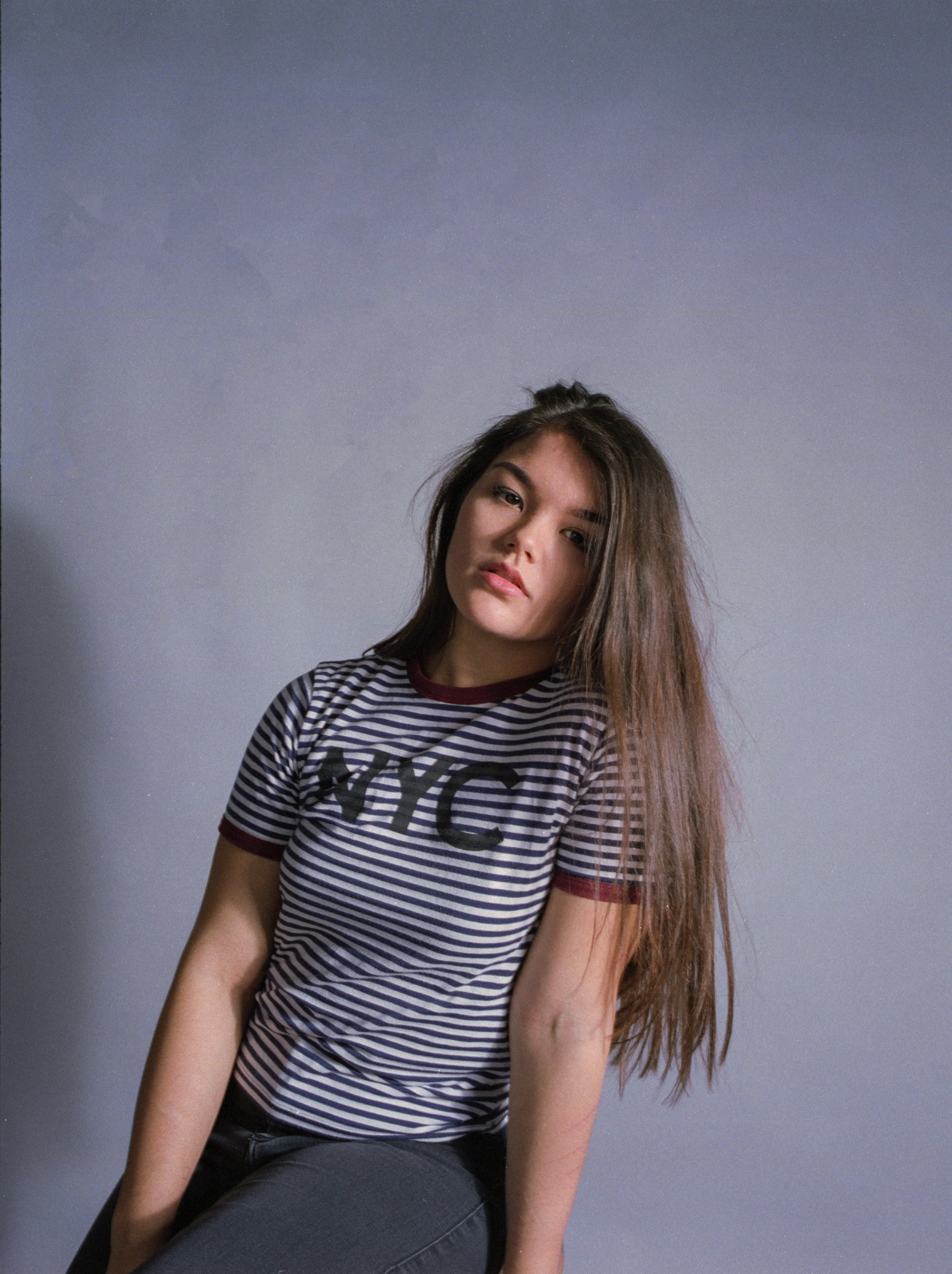 A young woman with long brown hair, wearing a striped NYC t-shirt, is sitting against a plain gray background, looking at the camera with a relaxed expression.
