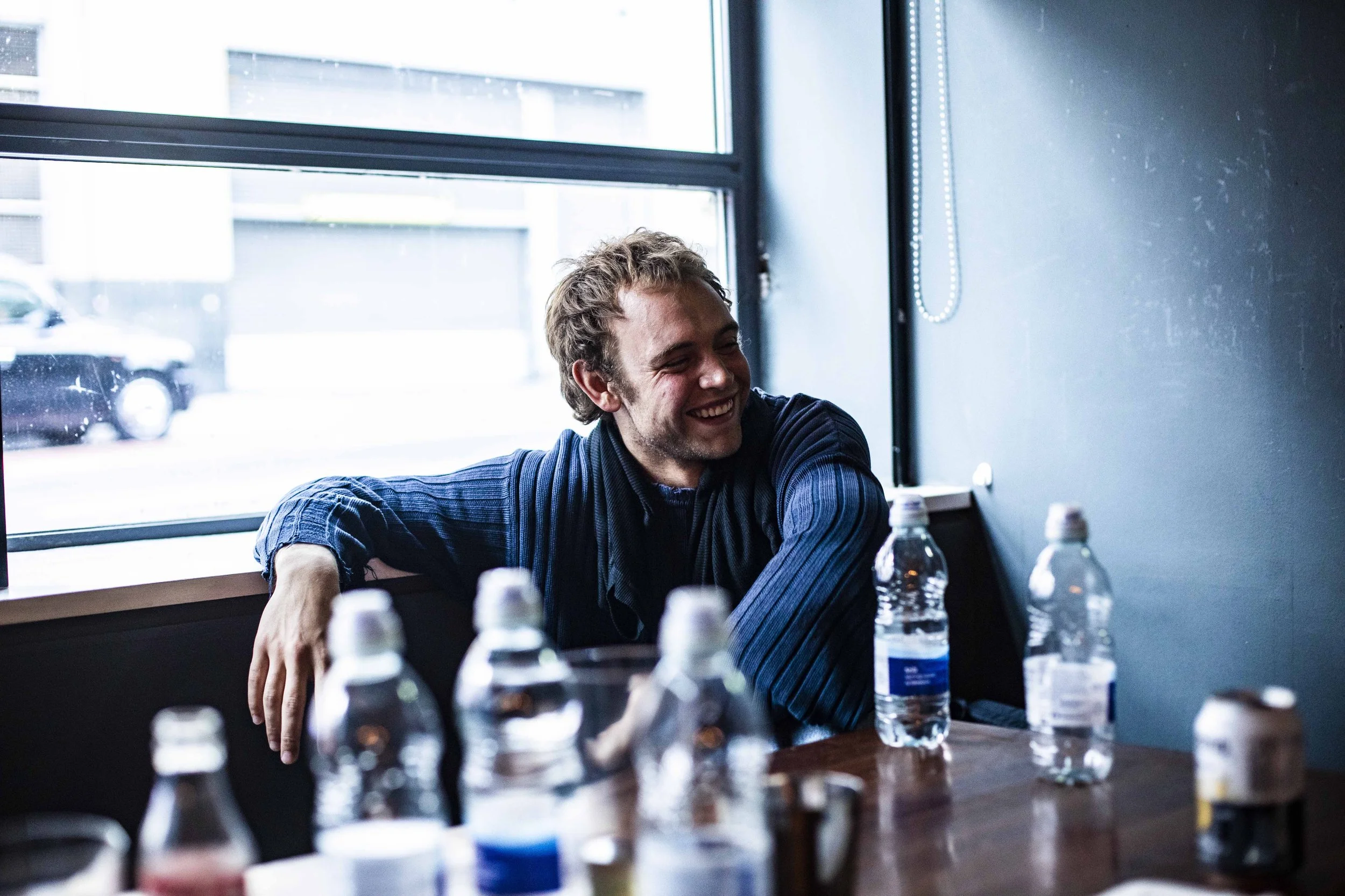 A young man with curly hair and a beard smiling and leaning on a table, surrounded by water bottles and plastic bottles, sitting near a large window in a room with blue walls.