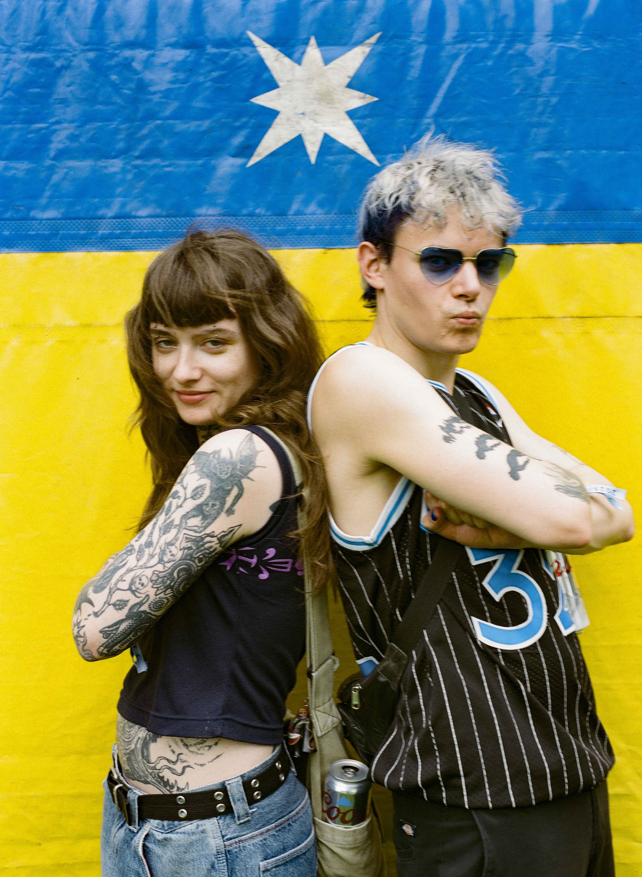 Two young people with tattoos posing back-to-back in front of a yellow and blue music festival with a white star decal above them.