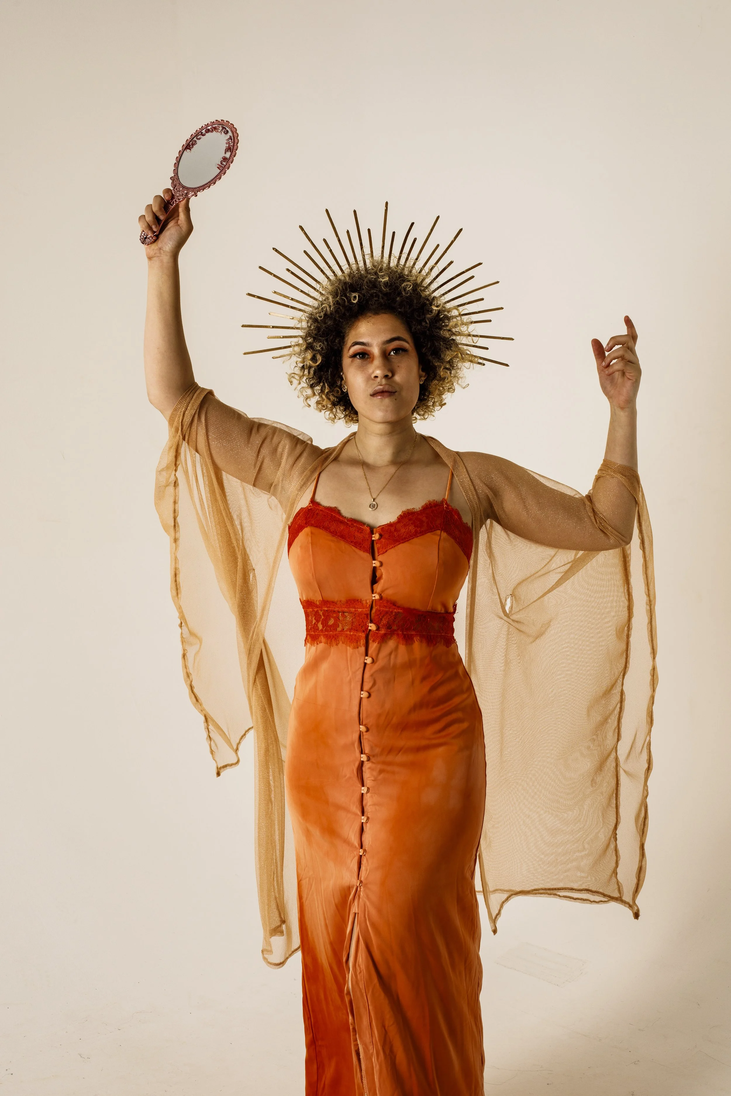 A woman with curly hair wearing an ornate gold halo headpiece, a sheer tan cover-up, and a form-fitting orange dress with red lace accents, holding a small hand mirror.