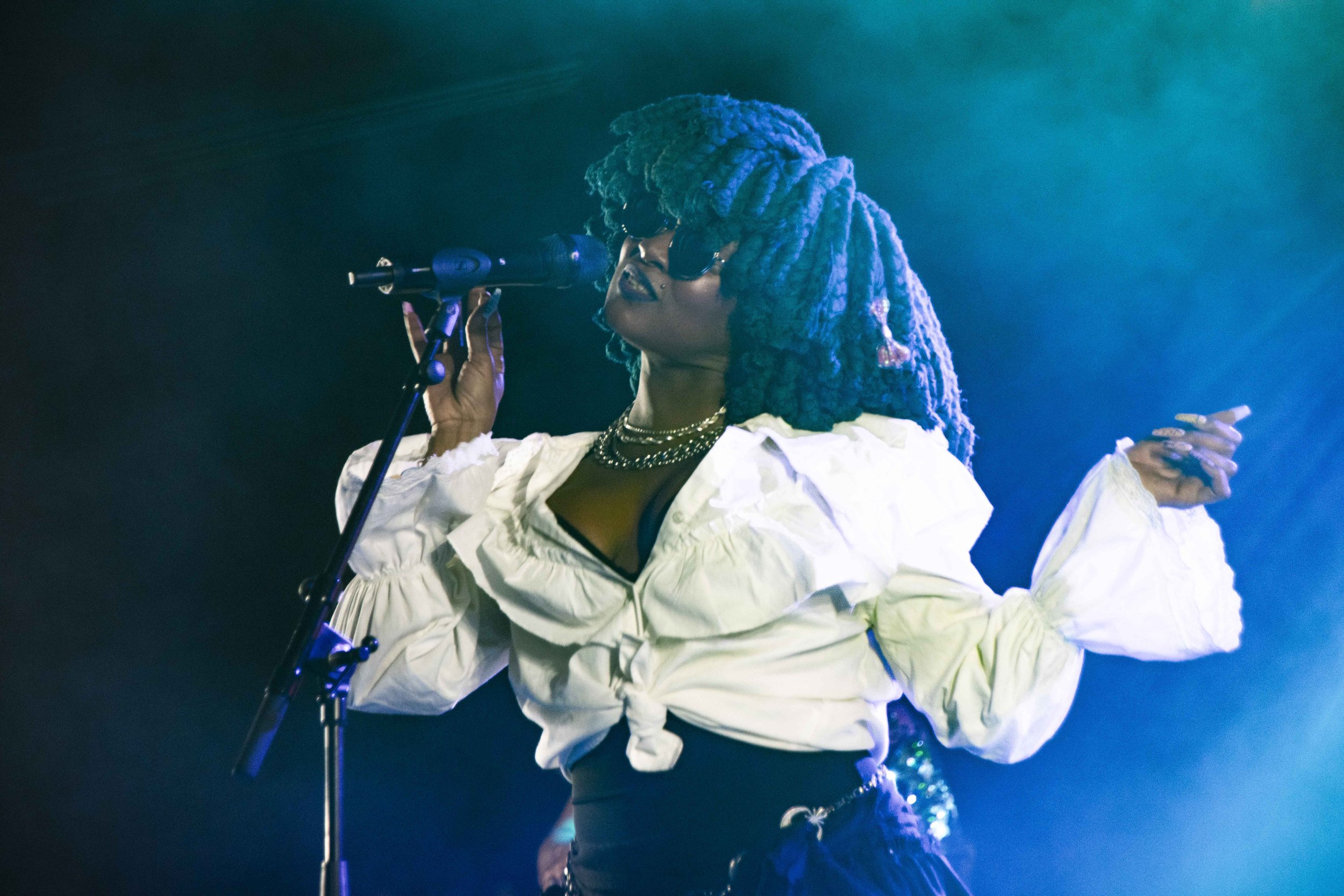 A female singer with blue dreadlocks and sunglasses performing on stage, holding a microphone, wearing a white tied-up blouse and layered necklaces, with stage lighting in the background.