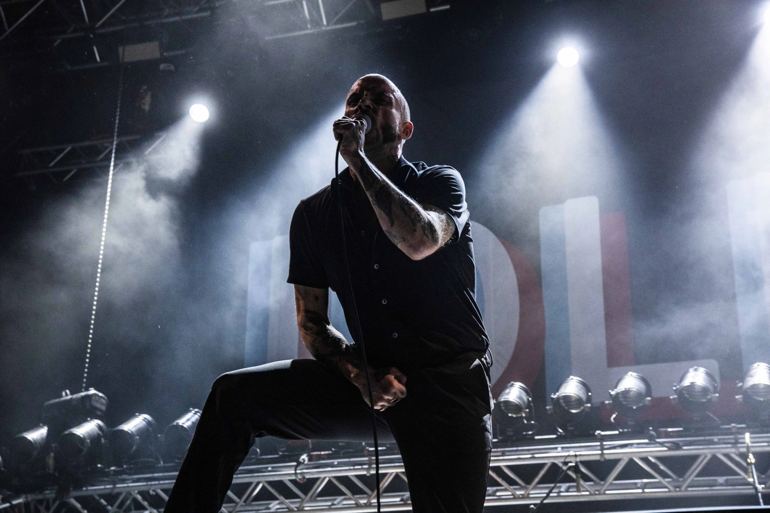 A male singer with tattoos on his arm, wearing a dark shirt, performs on stage with microphone in hand, backlit by stage lights in a concert setting.