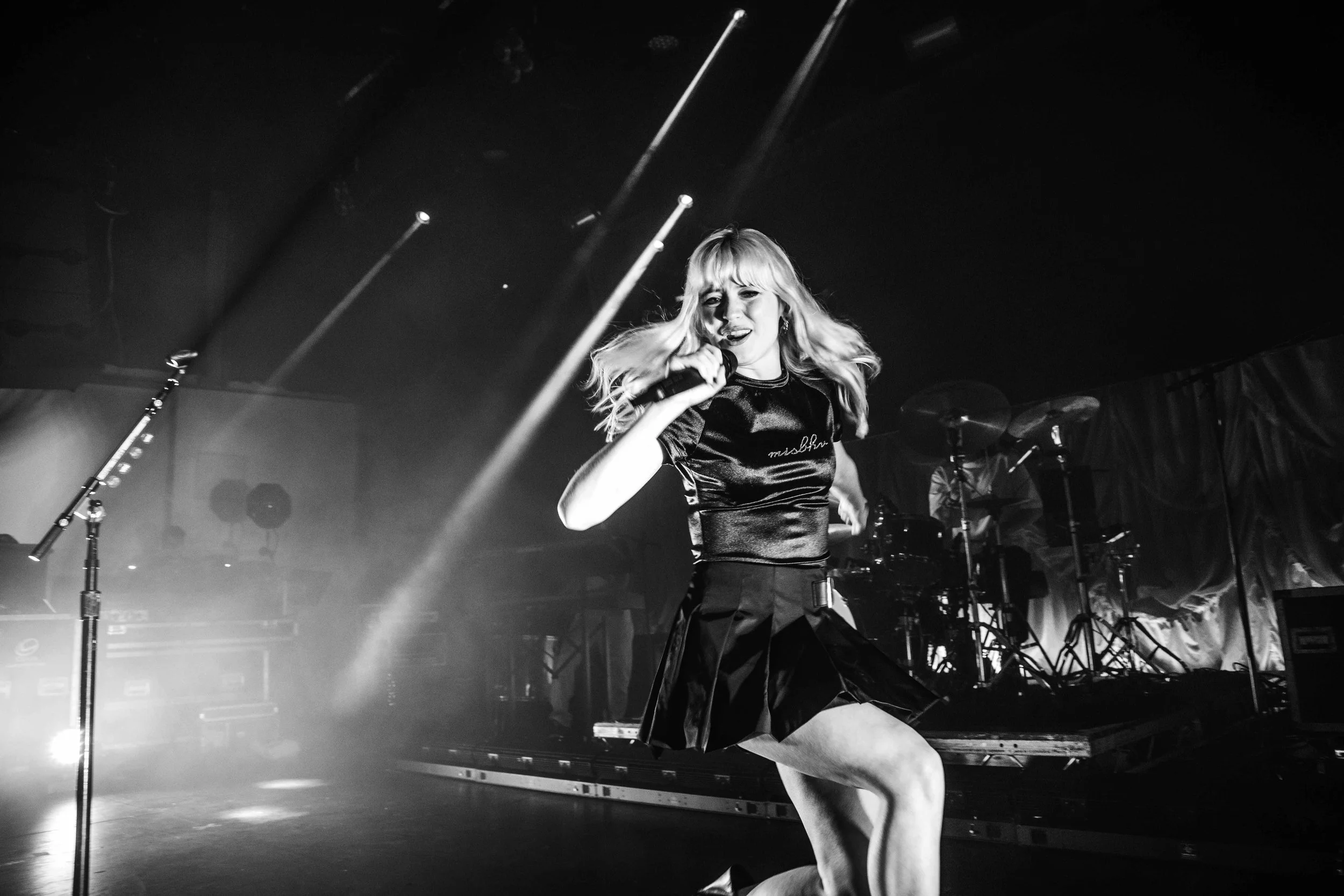 A female singer performing on stage, holding a microphone, with a band and drum set in the background, in a concert setting.