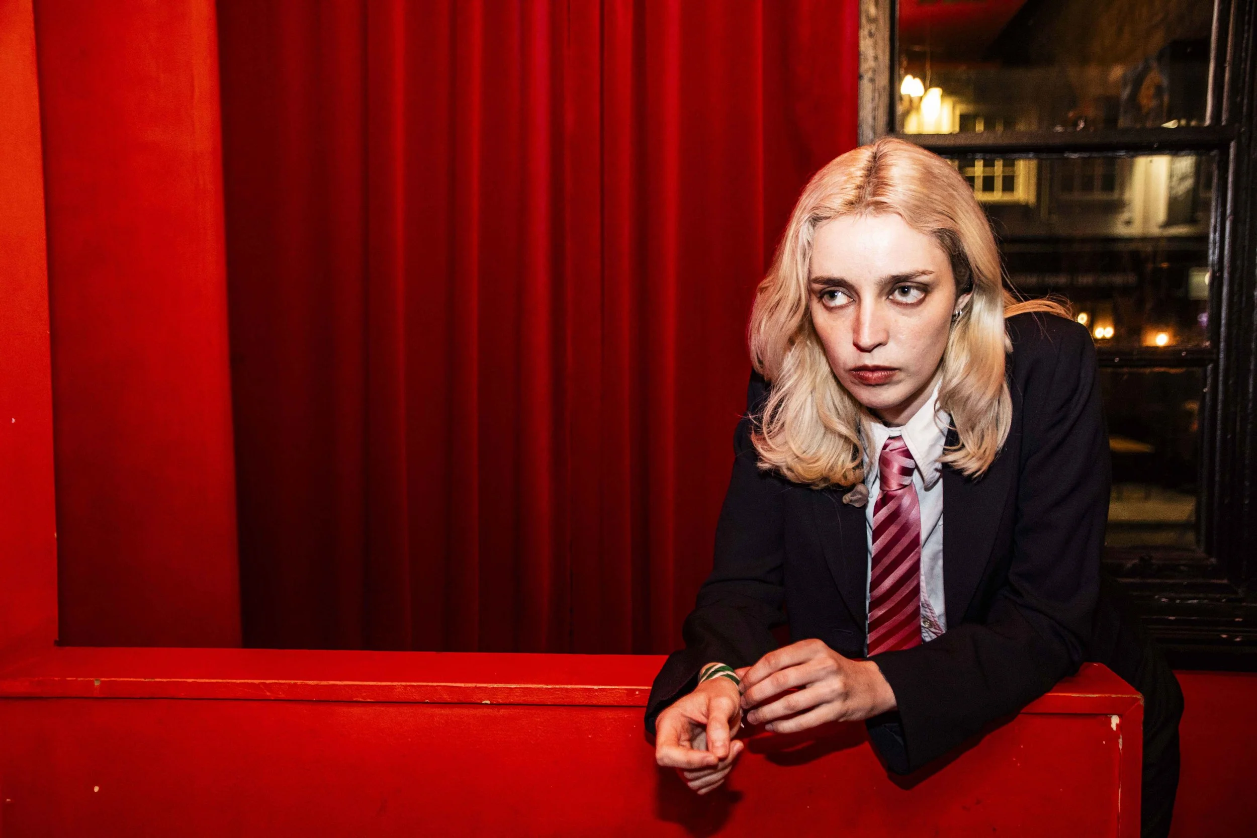 A woman with long blonde hair, dressed in a black blazer and striped tie, leaning on a red railing in a dimly lit room with red curtains and a window in the background.