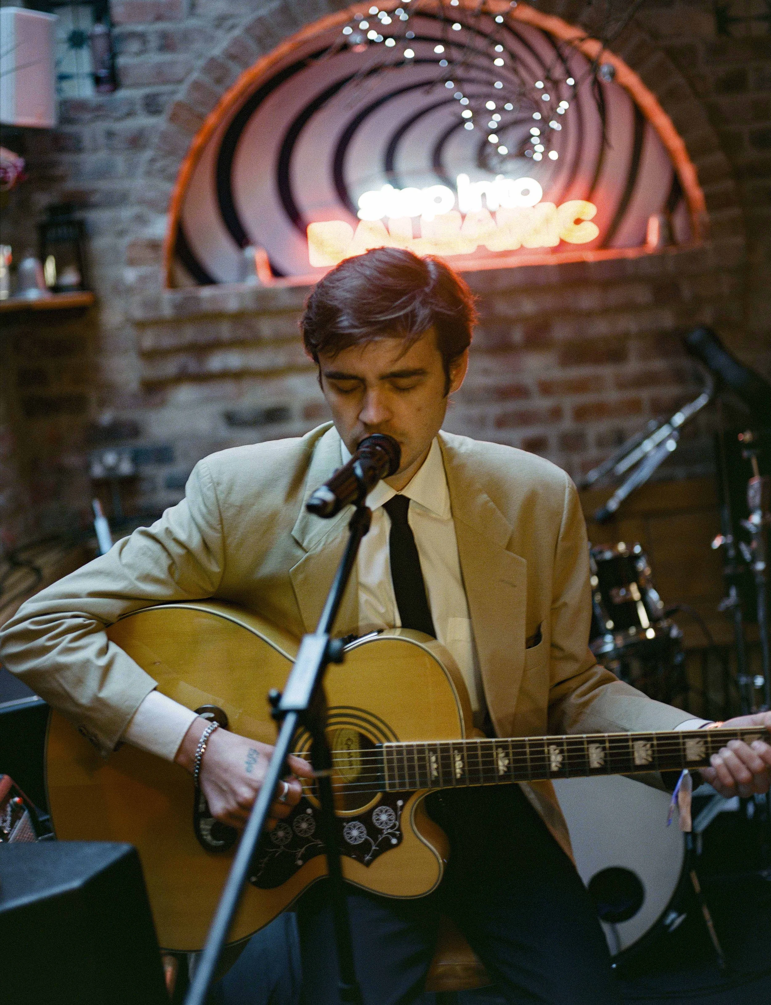 Young man in beige suit playing an acoustic guitar and singing into a microphone in a dimly lit bar with a brick wall background. Neon sign in the background reads 'Stay in the Darkness'.