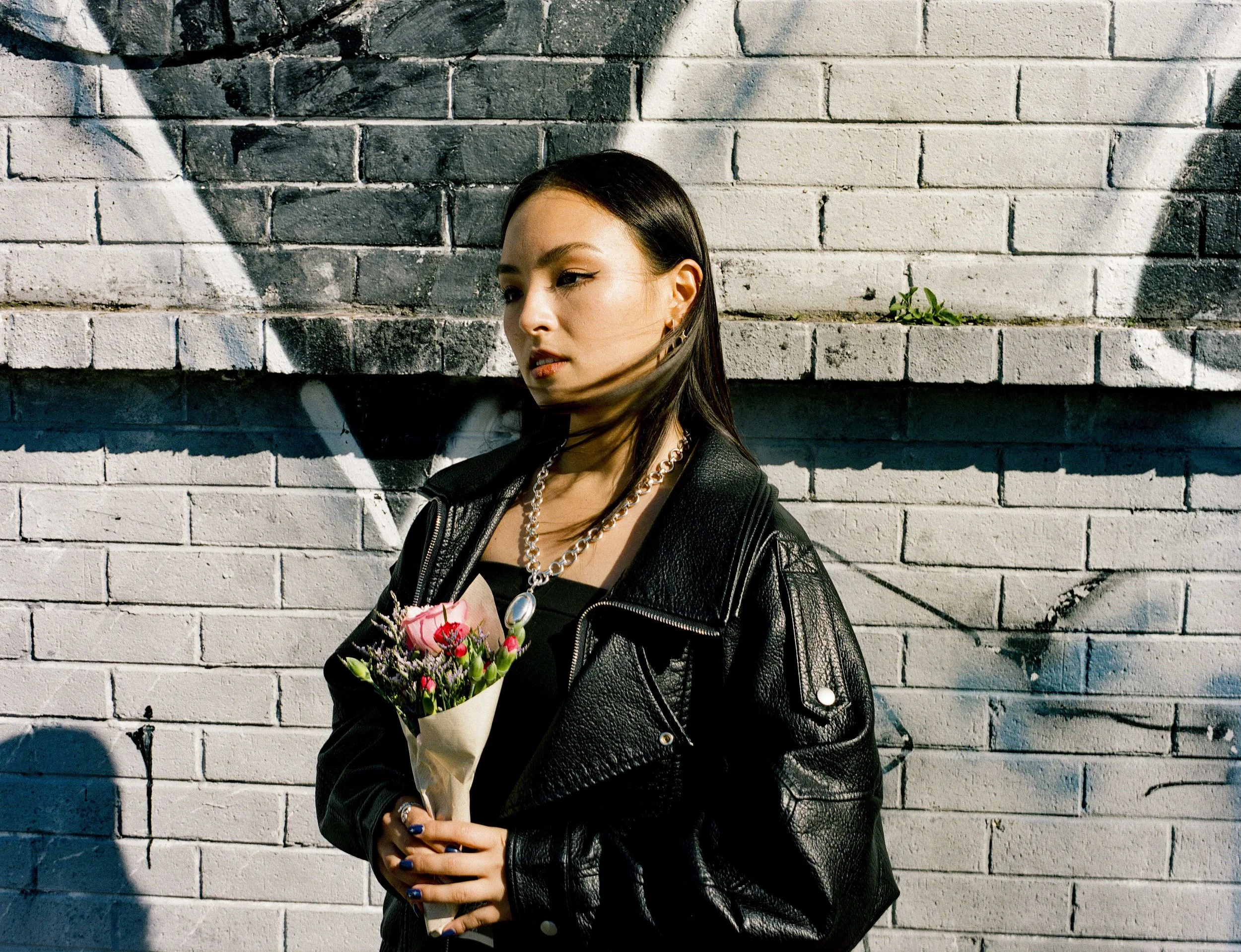 A woman standing in front of a white brick wall with graffiti, holding a bouquet of flowers, wearing a black leather jacket and a pearl necklace.