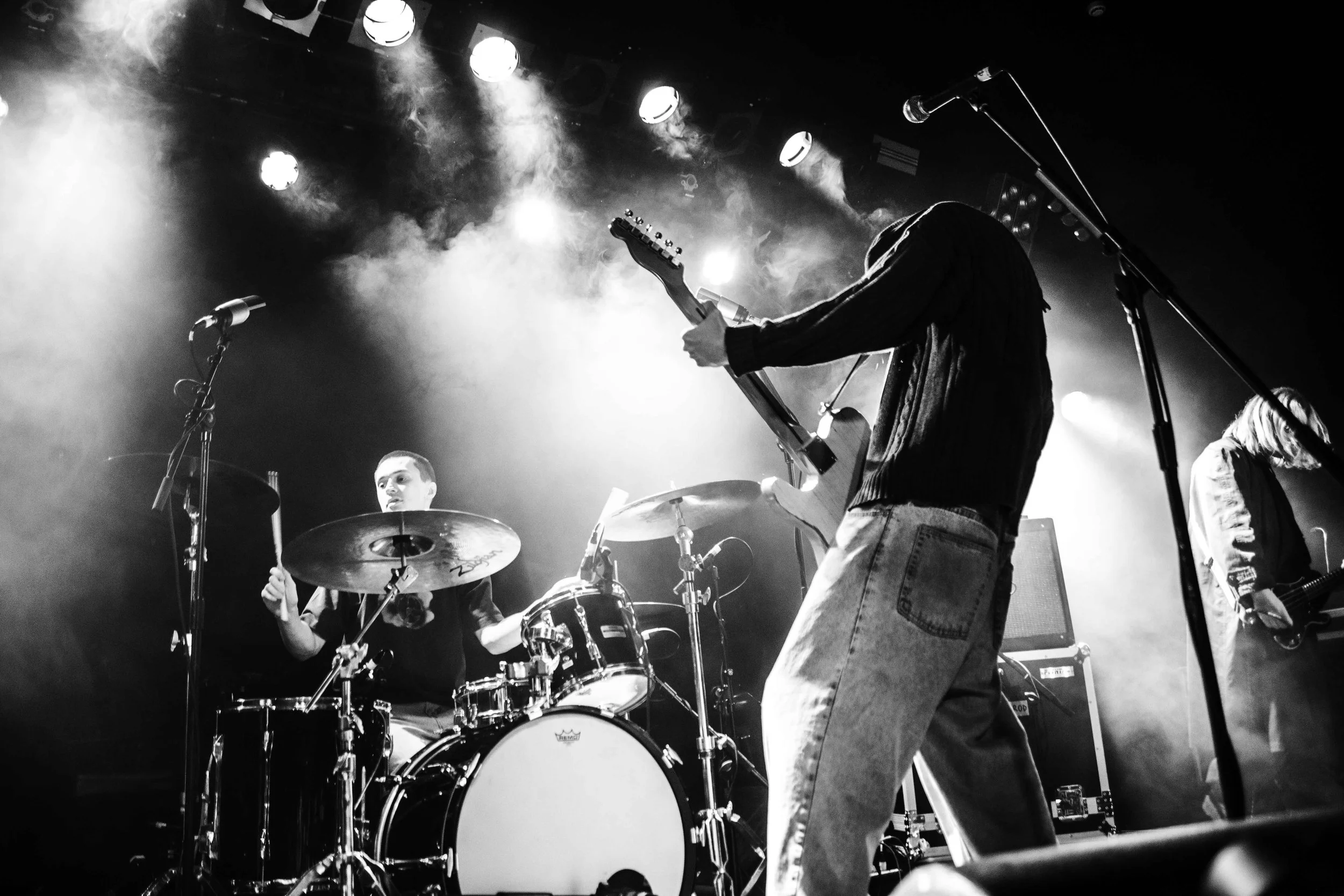 A black and white photo of a band performing on stage, with a drummer, a guitarist, and a bassist. The drummer is focused on playing, the bassist is shown from the side with long hair, and the guitarist is partially visible on the right. Stage lights