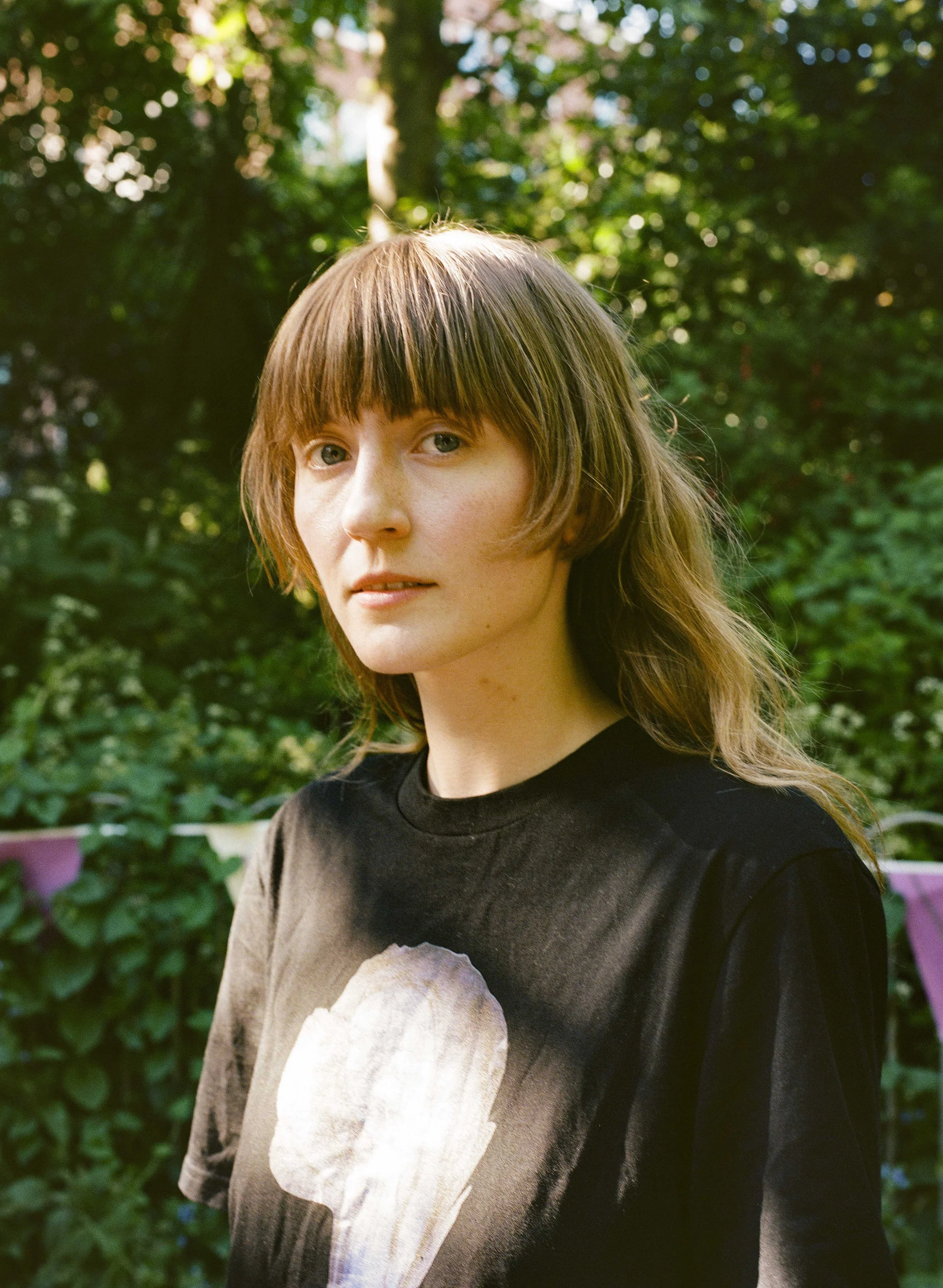 A young woman with shoulder-length brown hair and bangs, wearing a black t-shirt with a graphic of a seashell, standing outdoors with greenery in the background.