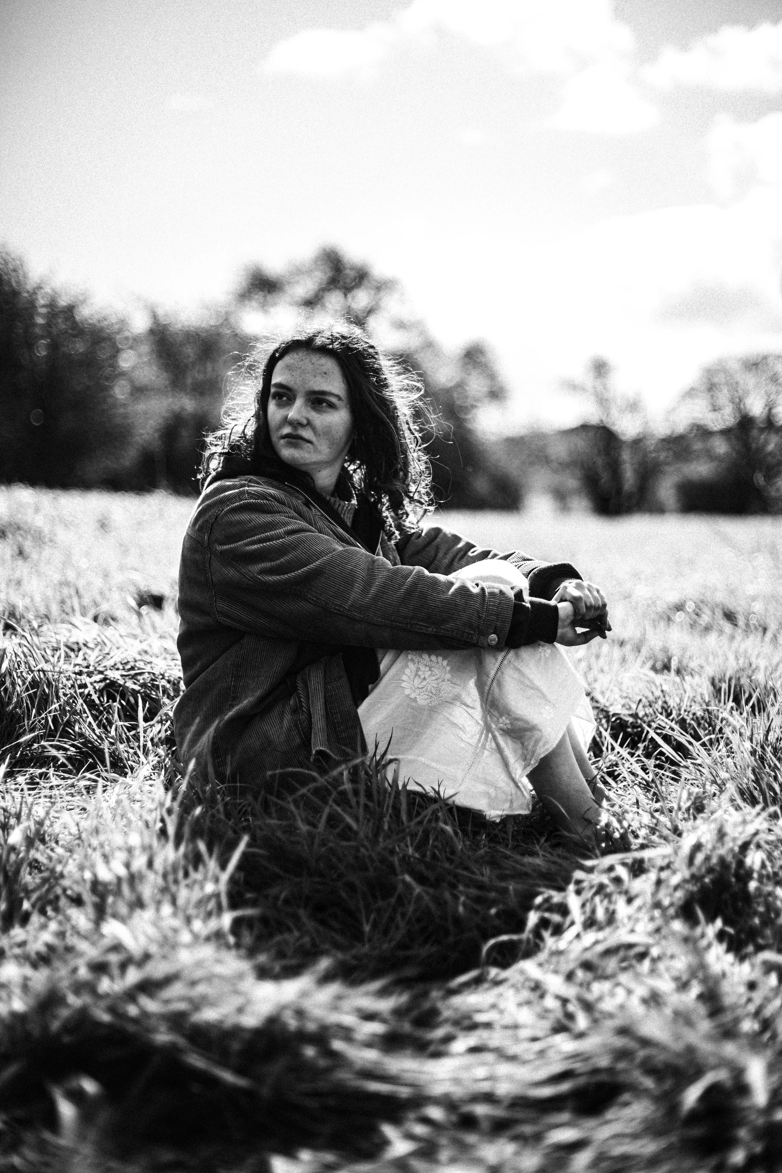 A woman sitting on grass outdoors in a field, looking to the side with a contemplative expression, black and white photograph.