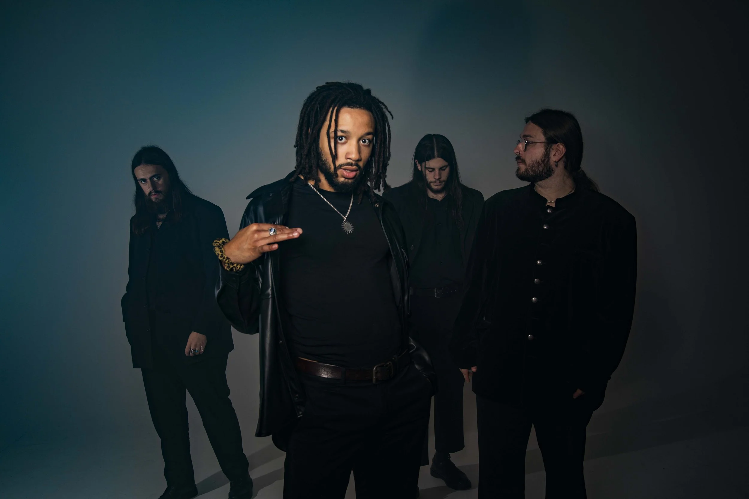 Four men with dark clothing and long hair posing against a dark background, with one man in the front gesturing with his hand.