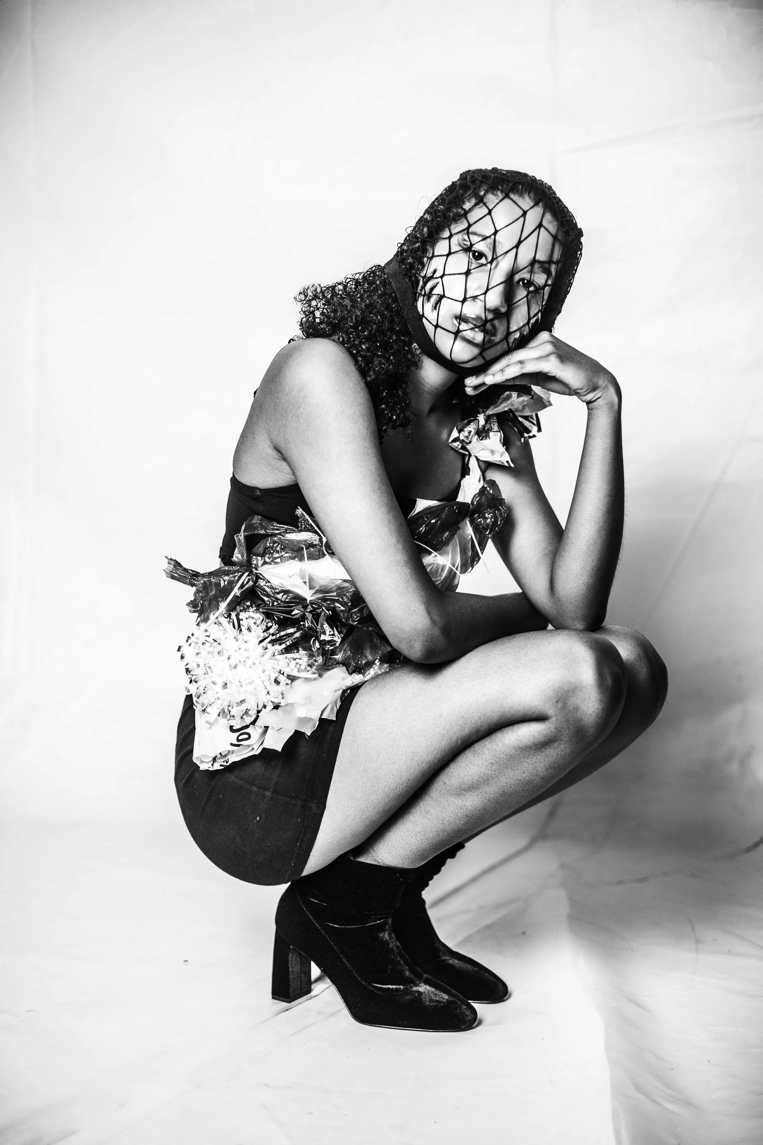 Black and white photo of a woman crouching, wearing a netted face mask and a dress made of crumpled paper and foil, paired with ankle boots.