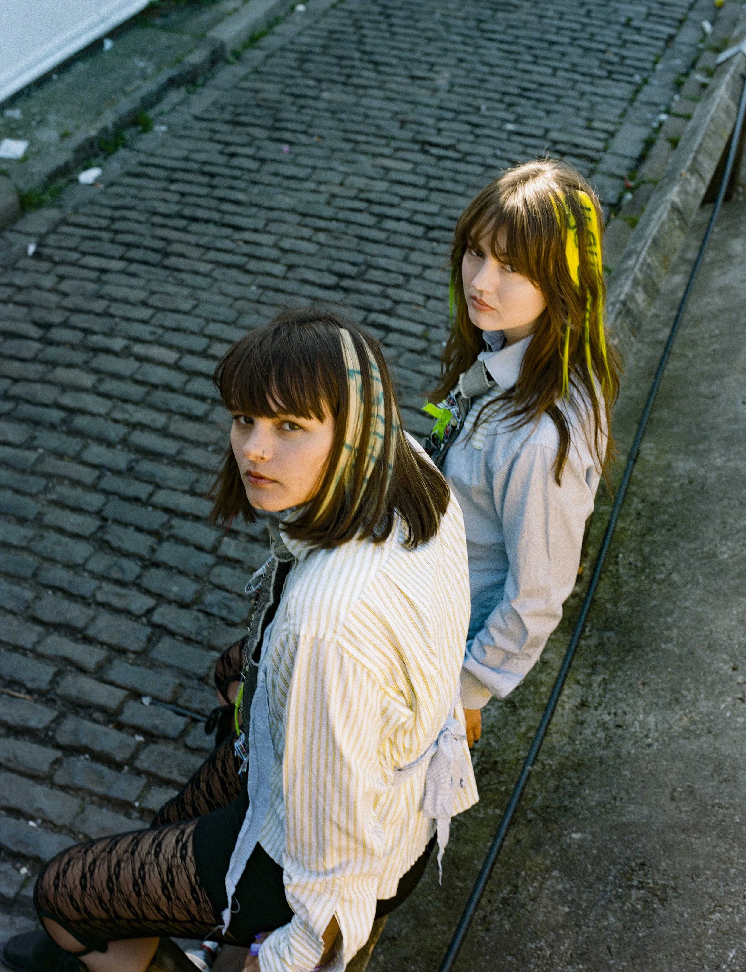 Two young women with colorful streaks in their hair, sitting on a bench on a cobblestone street, looking up at the camera.