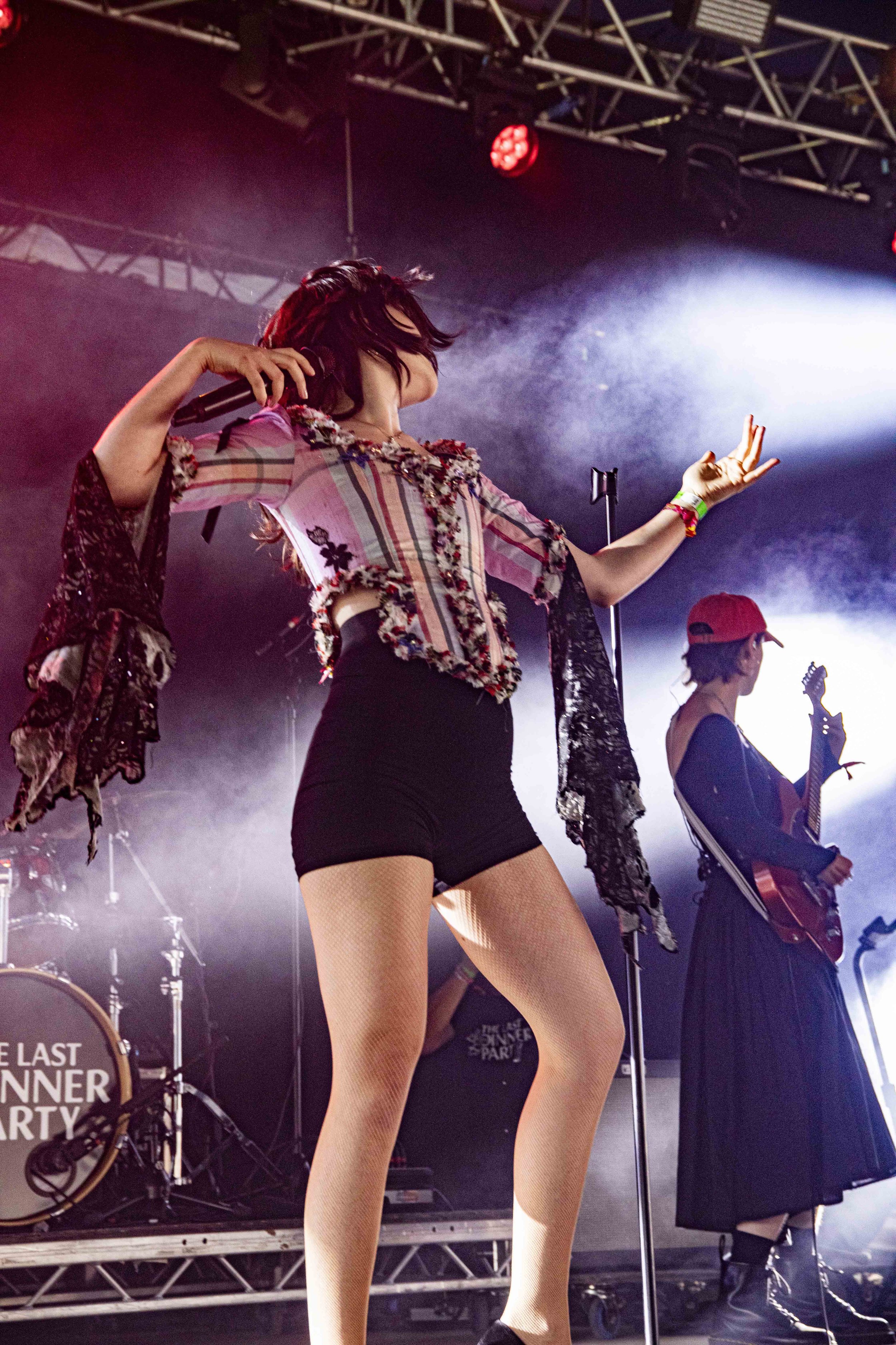 Female singer performing on stage with a microphone, wearing a floral-patterned blouse, shorts, and fishnet stockings, with guitarist in the background.
