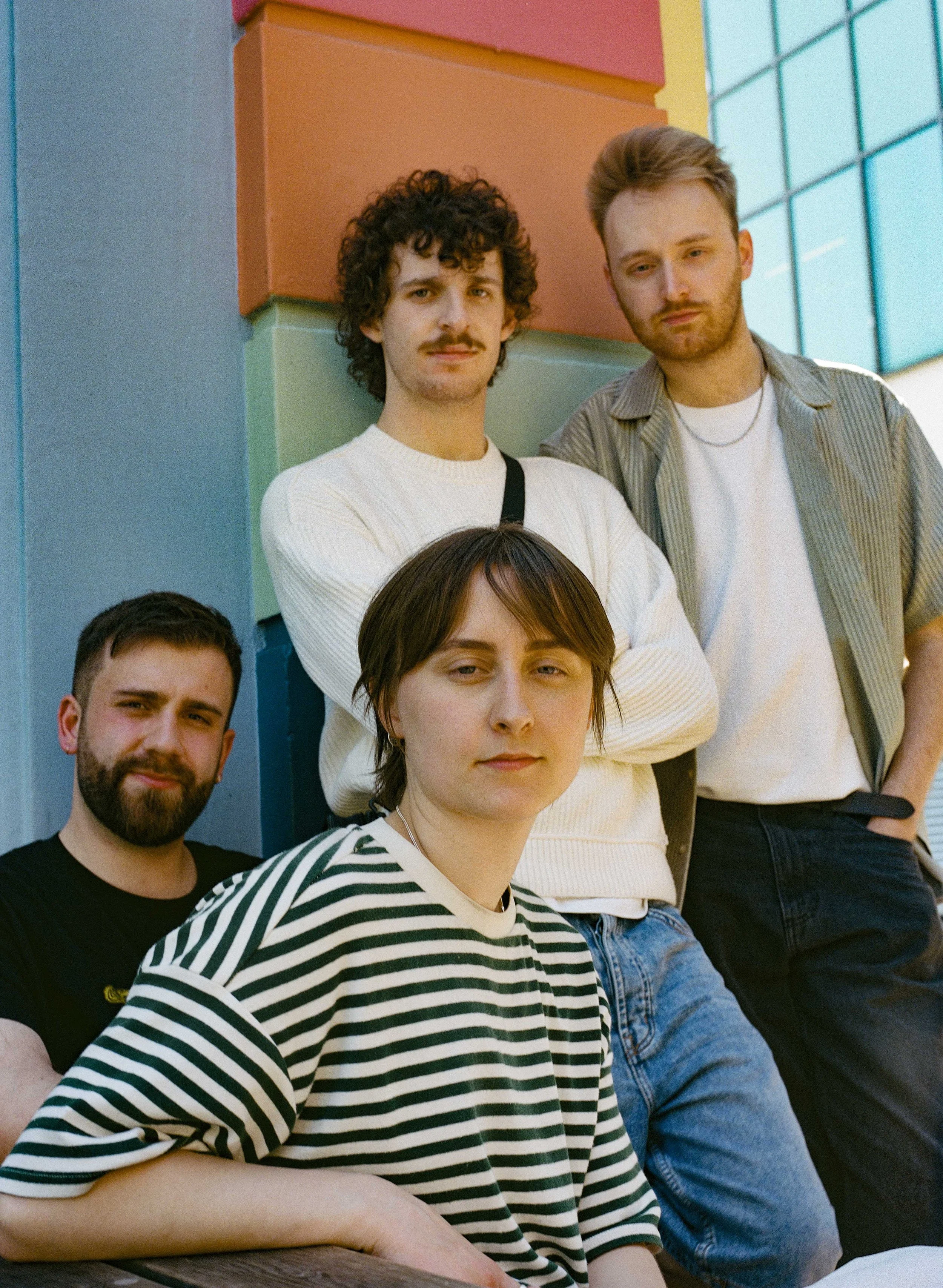A group of five young adults, 4 men and 1 woman, posing outdoors against a colourful wall and building, with casual clothing and relaxed expressions.