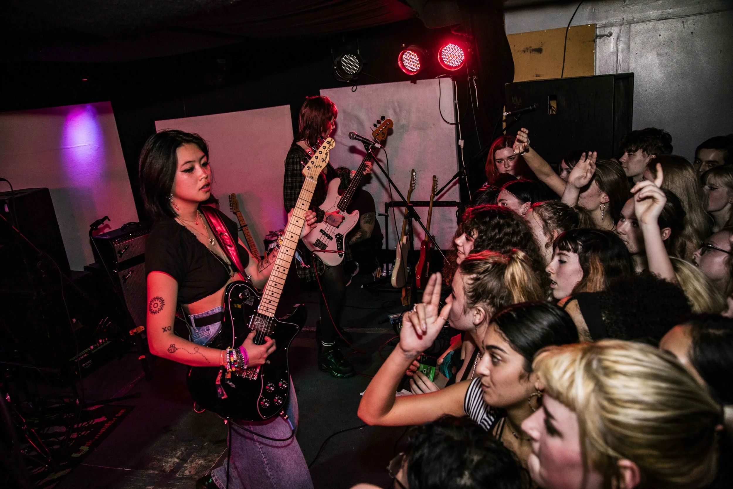 A live concert scene with two female musicians on stage. One is playing an electric guitar, and the other is singing and playing an instrument. The audience is close to the stage, engaged and enjoying the performance.