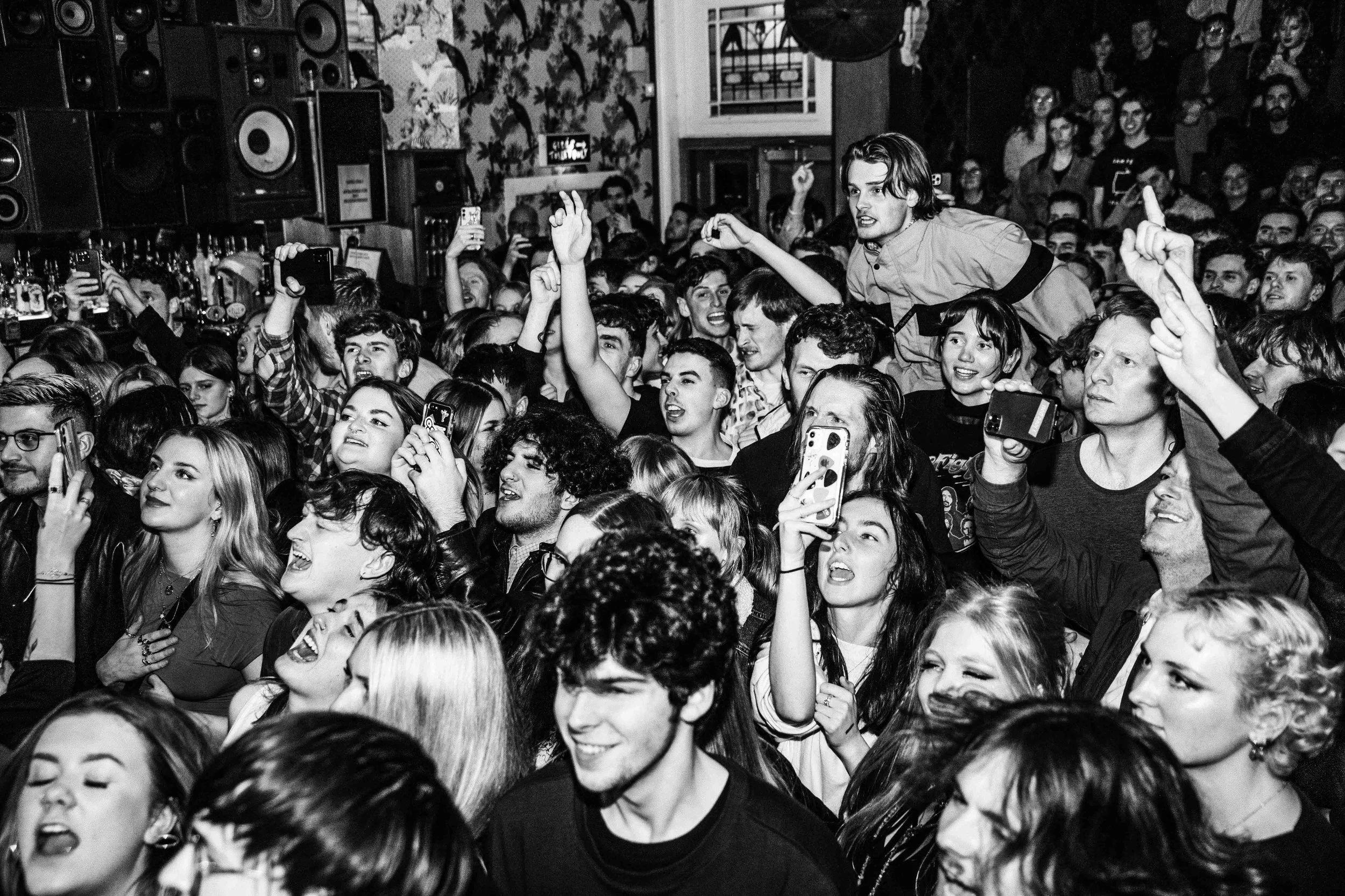 Crowd of young people at a concert or event, with some taking photos and others cheering. A person is being held above the crowd.