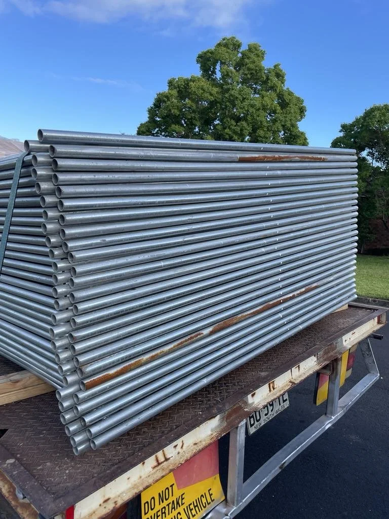 A large stack of metal pipes on the bed of a flatbed truck outdoors with trees and blue sky in the background.