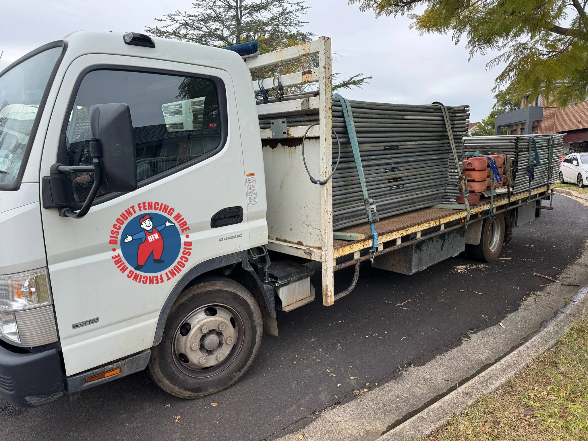 A white utility truck parked on the street with a logo that says 'Discount Fencing Hire' on the door. The truck's flatbed is loaded with metal fencing material, some orange safety equipment, and black pipes secured with straps.