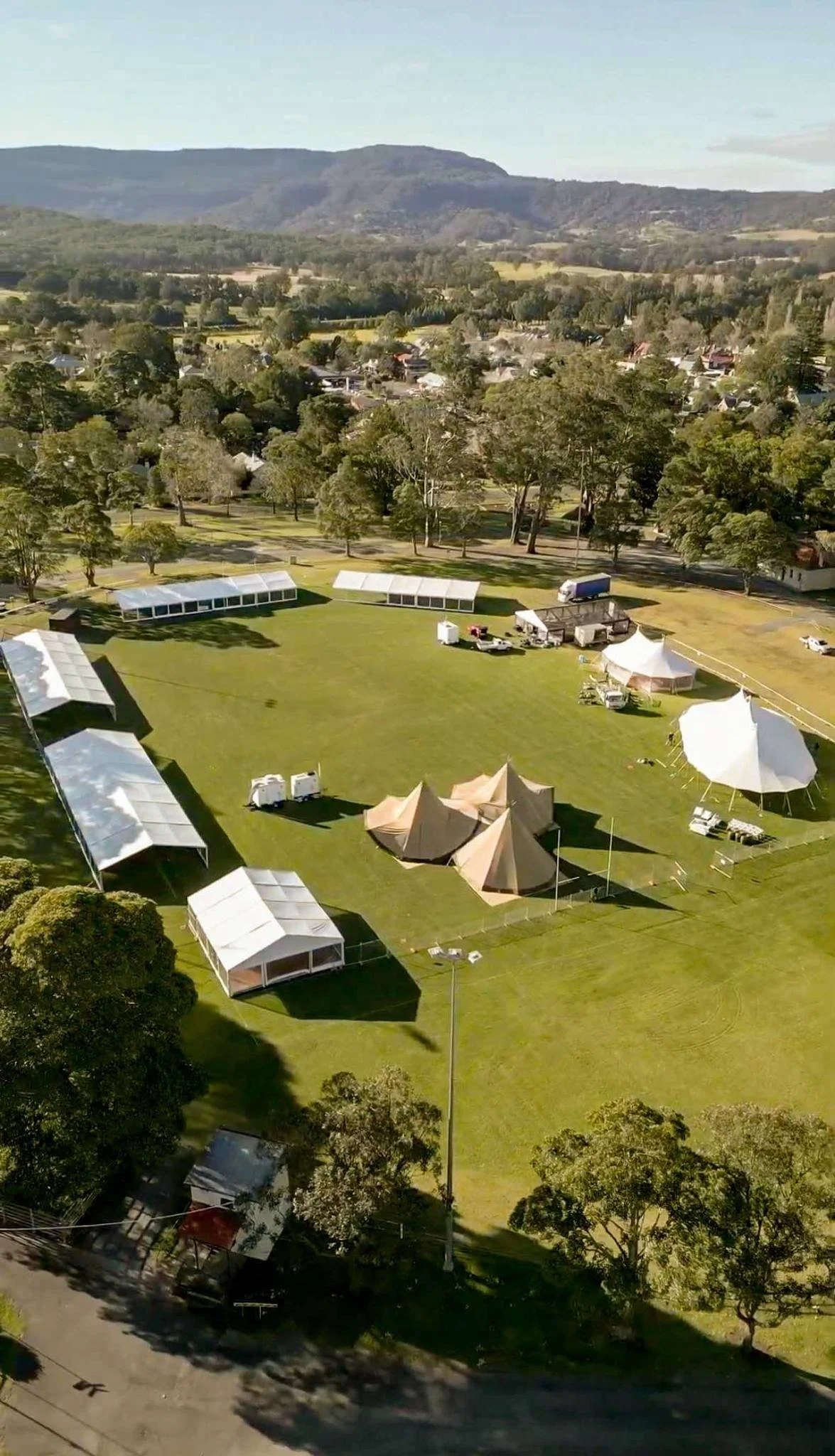 An aerial view of a grassy park area with multiple white tents and structures, some being large event tents, set up for a festival or event in a scenic location with trees, roads, and hills in the background.