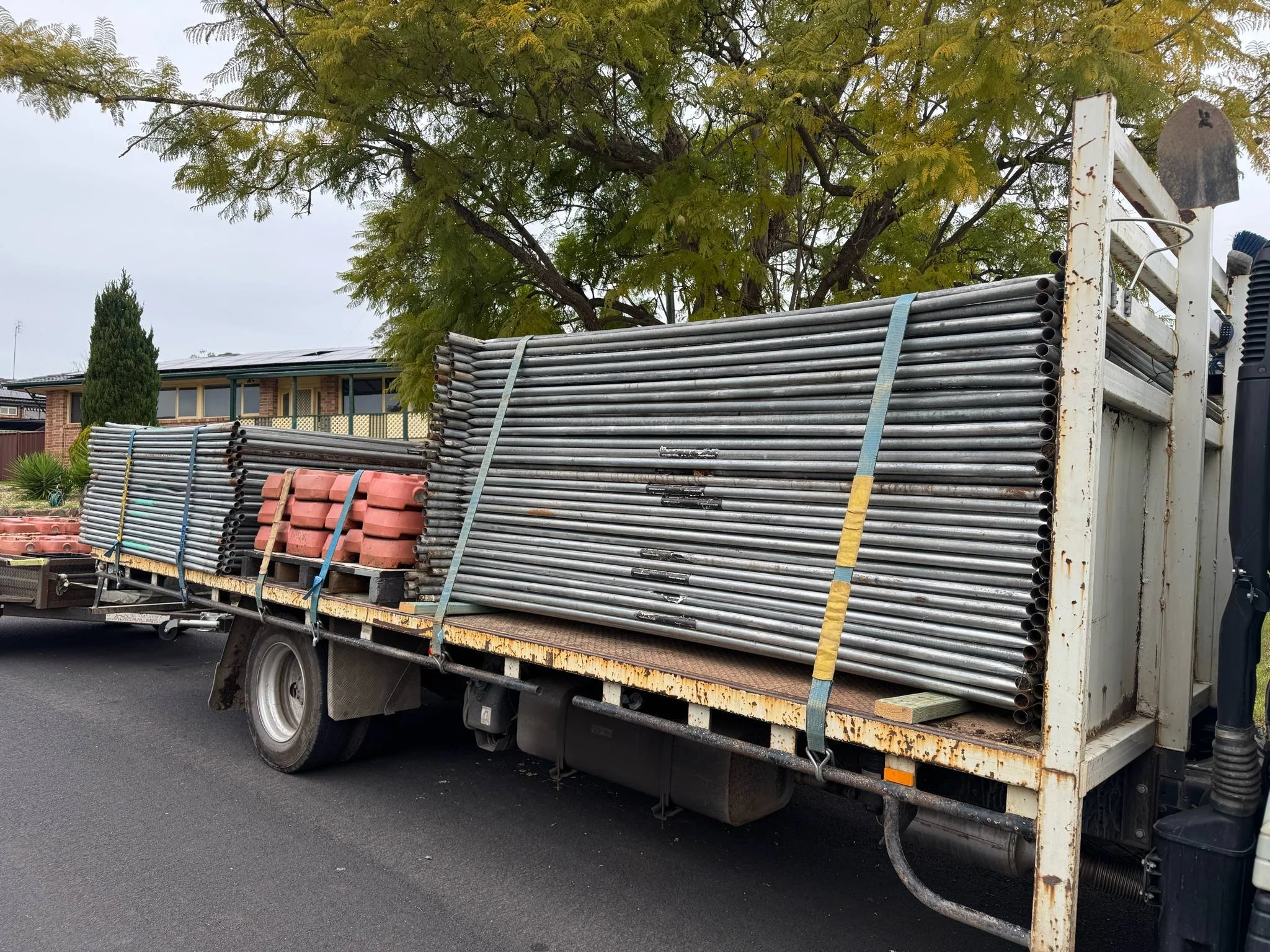 Flatbed truck carrying metal pipes and concrete blocks parked on suburban street with trees and house in the background.