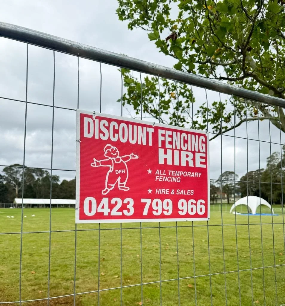 Red and white sign attached to a metal fence advertising discount fencing hire with a cartoon character and contact number, set against a background of a grassy field and cloudy sky.
