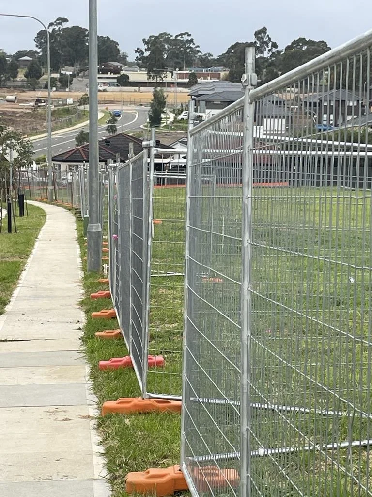 Fencing barriers with orange bases set up along a sidewalk in a residential area, with houses, trees, and a street in the background.