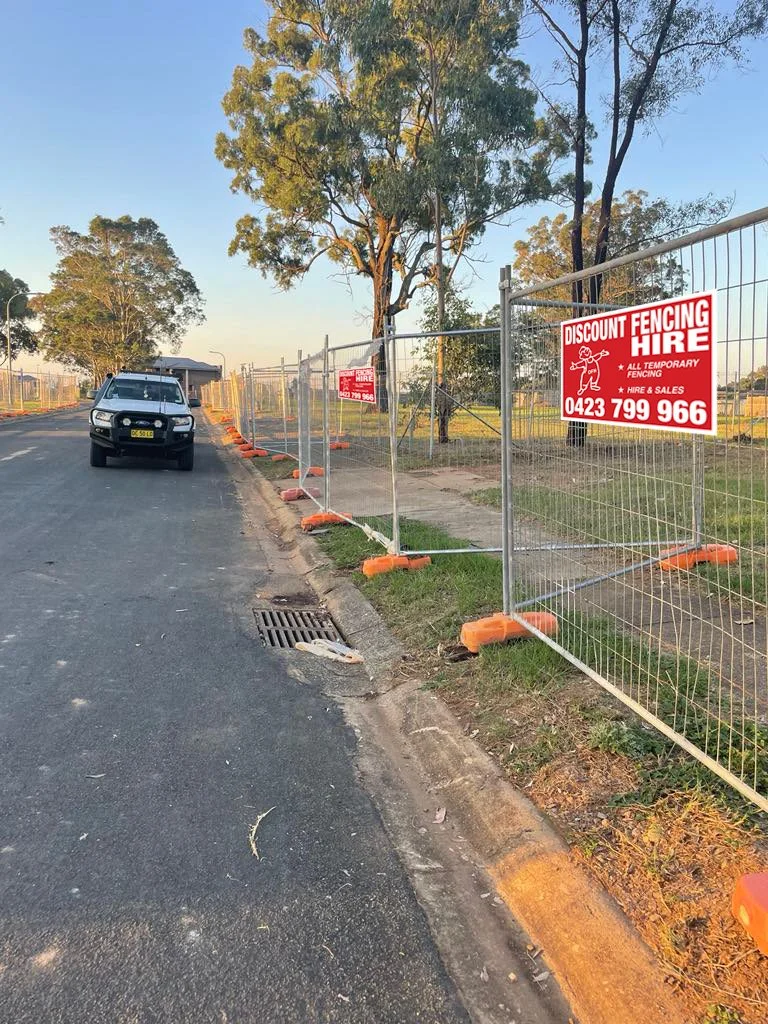 Street scene with a parked white vehicle, construction fencing with signs for fencing hire, trees in the background, and a clear sky.