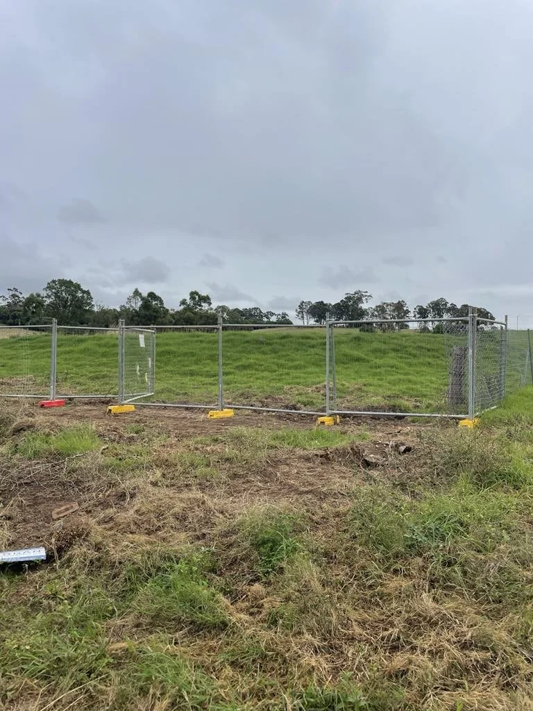 A chain-link fence surrounds a grassy hilltop under a cloudy sky, with construction barriers at the fence base.