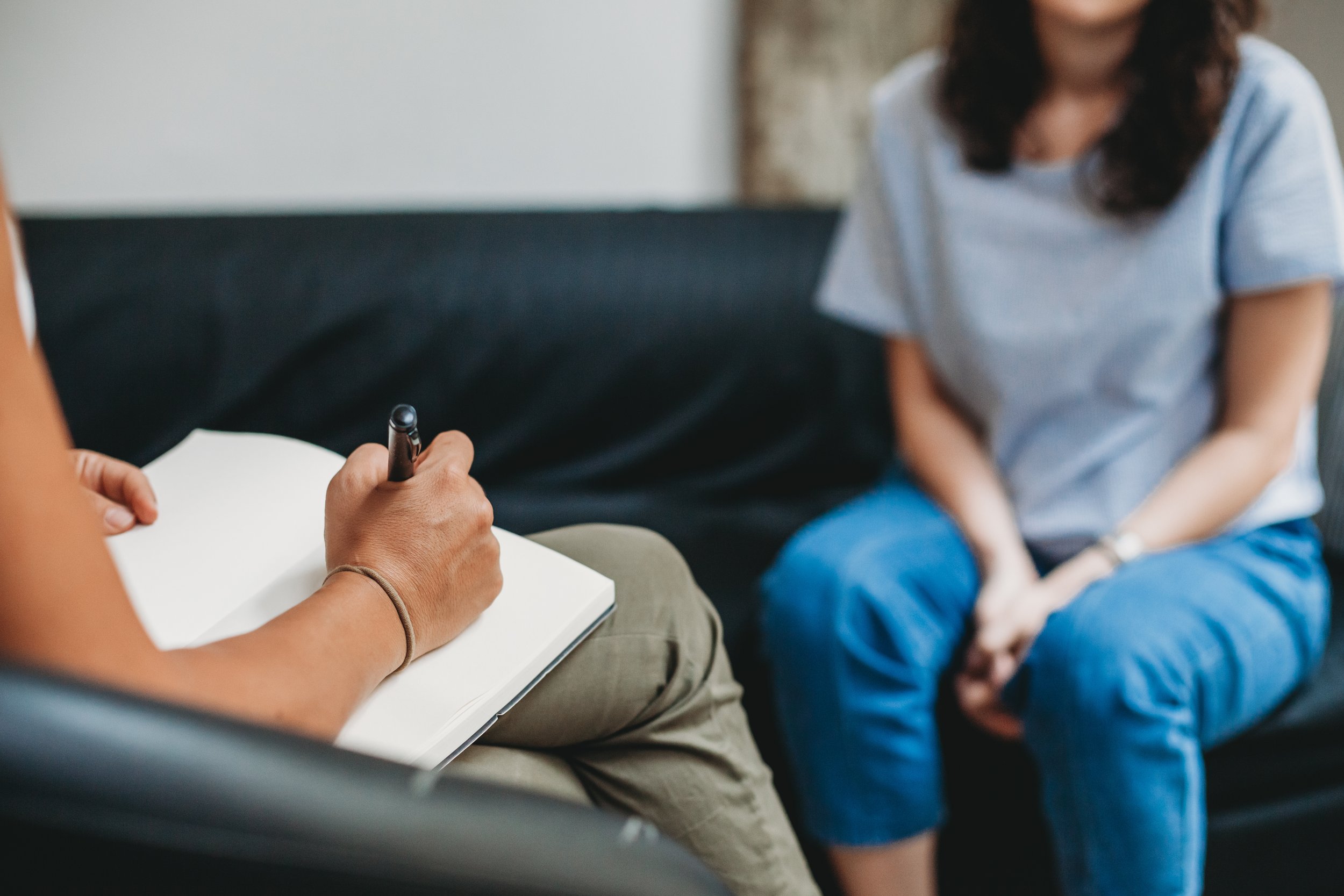Person taking notes during a therapy session with another person sitting on a couch.