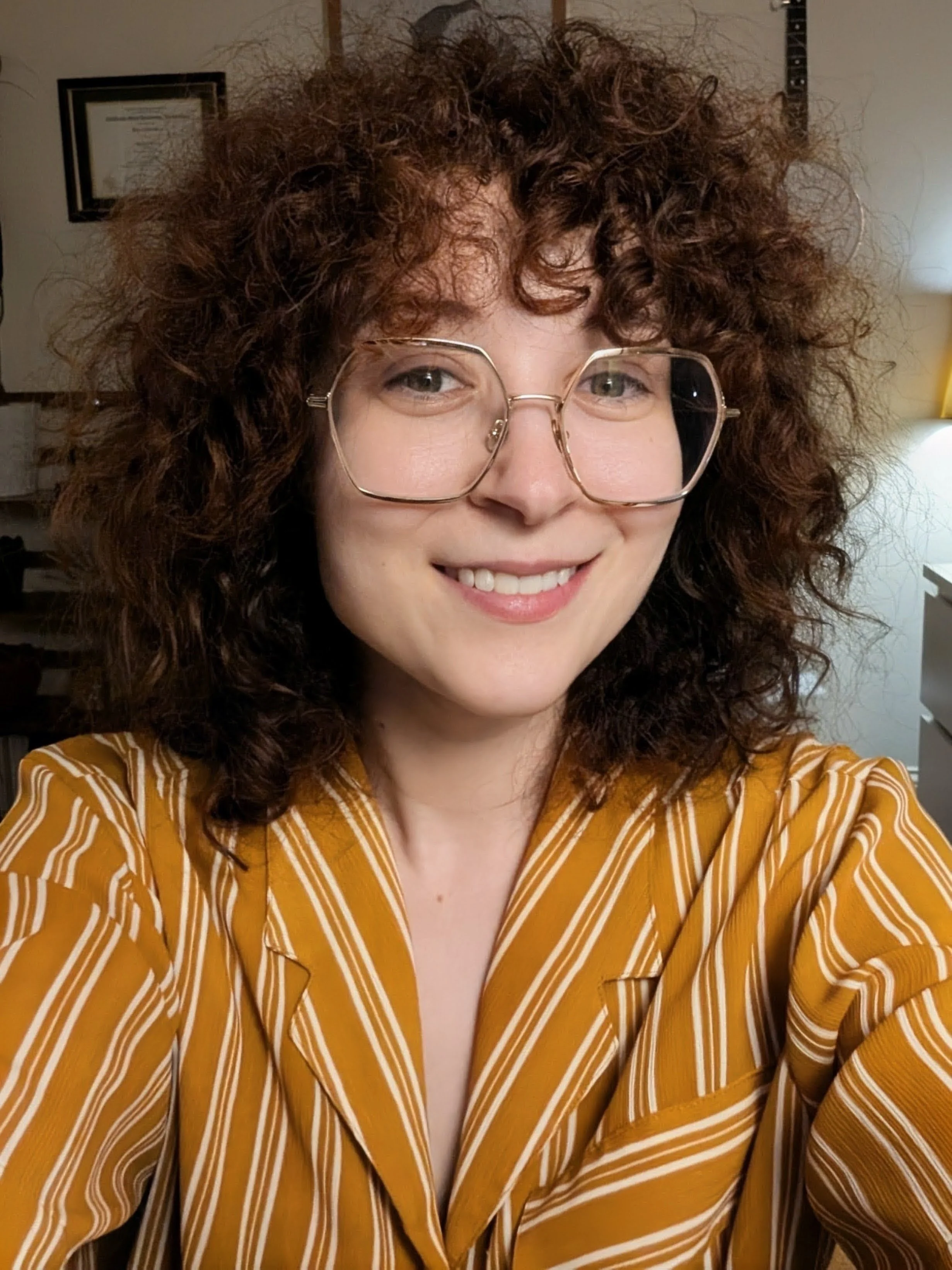 A woman with curly hair and glasses smiling, wearing a yellow striped blouse in an indoor setting.