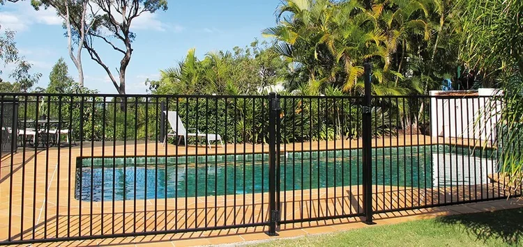 Residential backyard with a swimming pool, surrounded by a black metal fence, lounge chairs, and lush green trees.