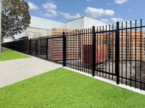 Black metal fence along sidewalk with green grass and construction site with brick walls and buildings in background.